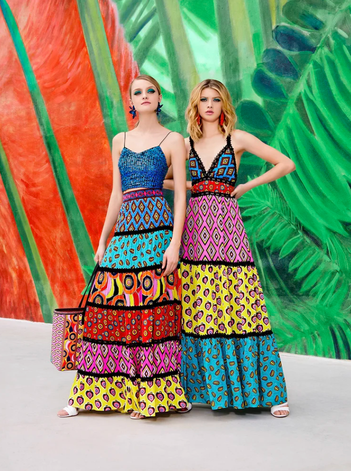 Two women in colorful, patterned maxi dresses standing in front of a vibrant green and orange tropical mural.