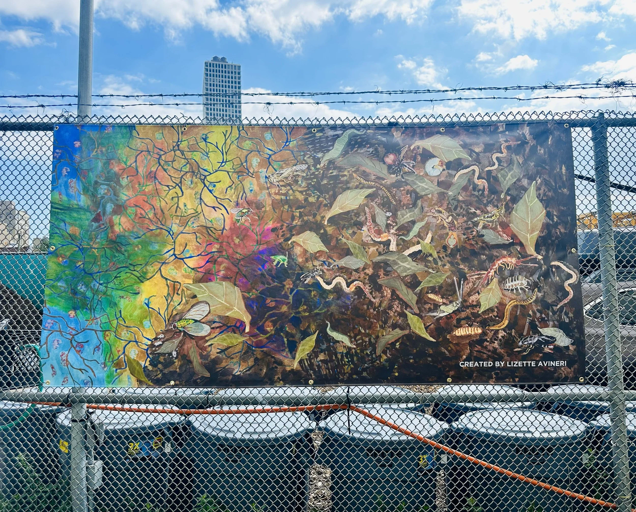 Colorful outdoor painting of insects and plants behind a chain-link fence and barbed wire, with a city skyline in the background.