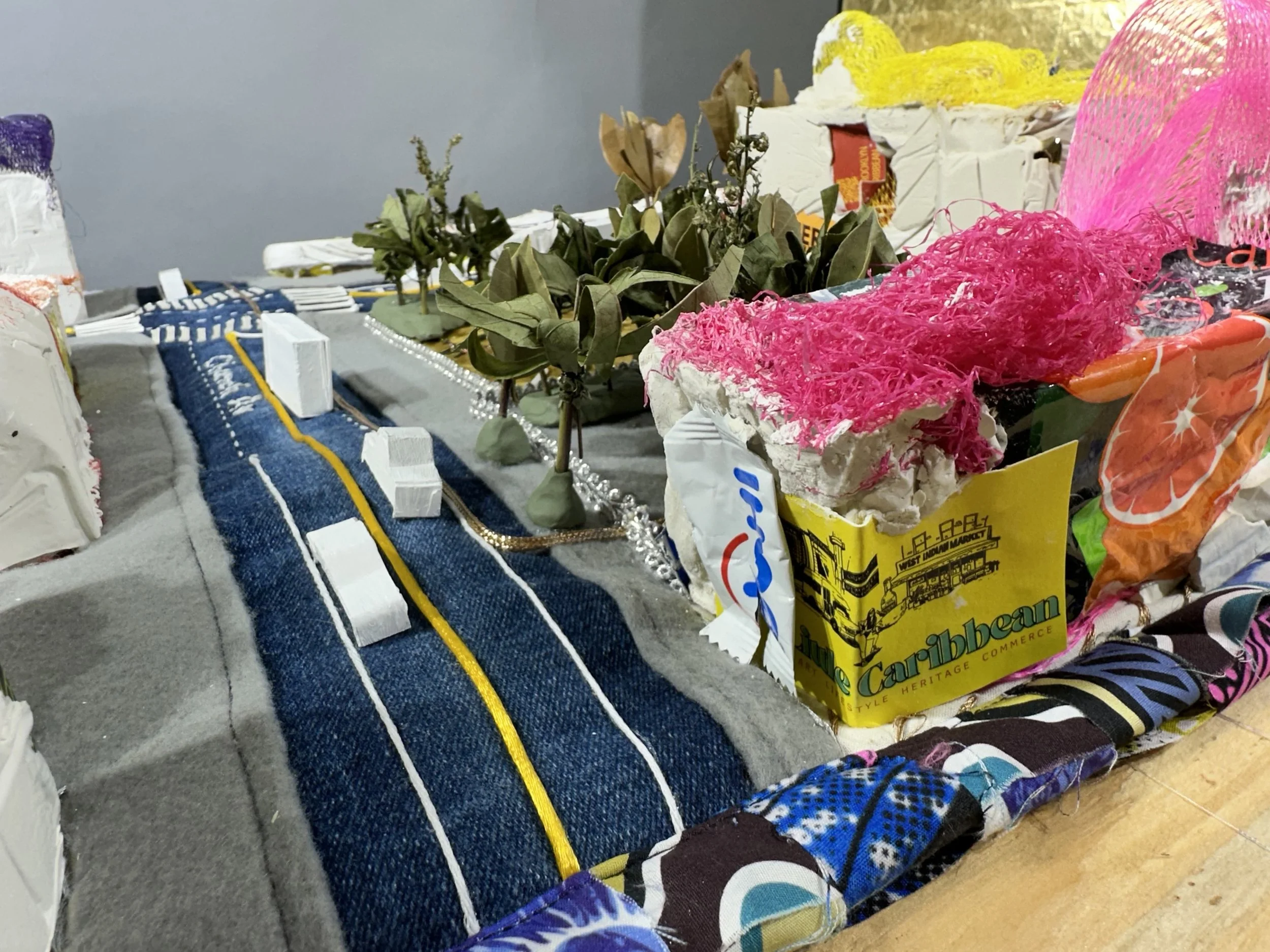 A model of a city street scene made with various small objects, including fabric, paper, and natural elements like leaves, representing roads, trees, and buildings.