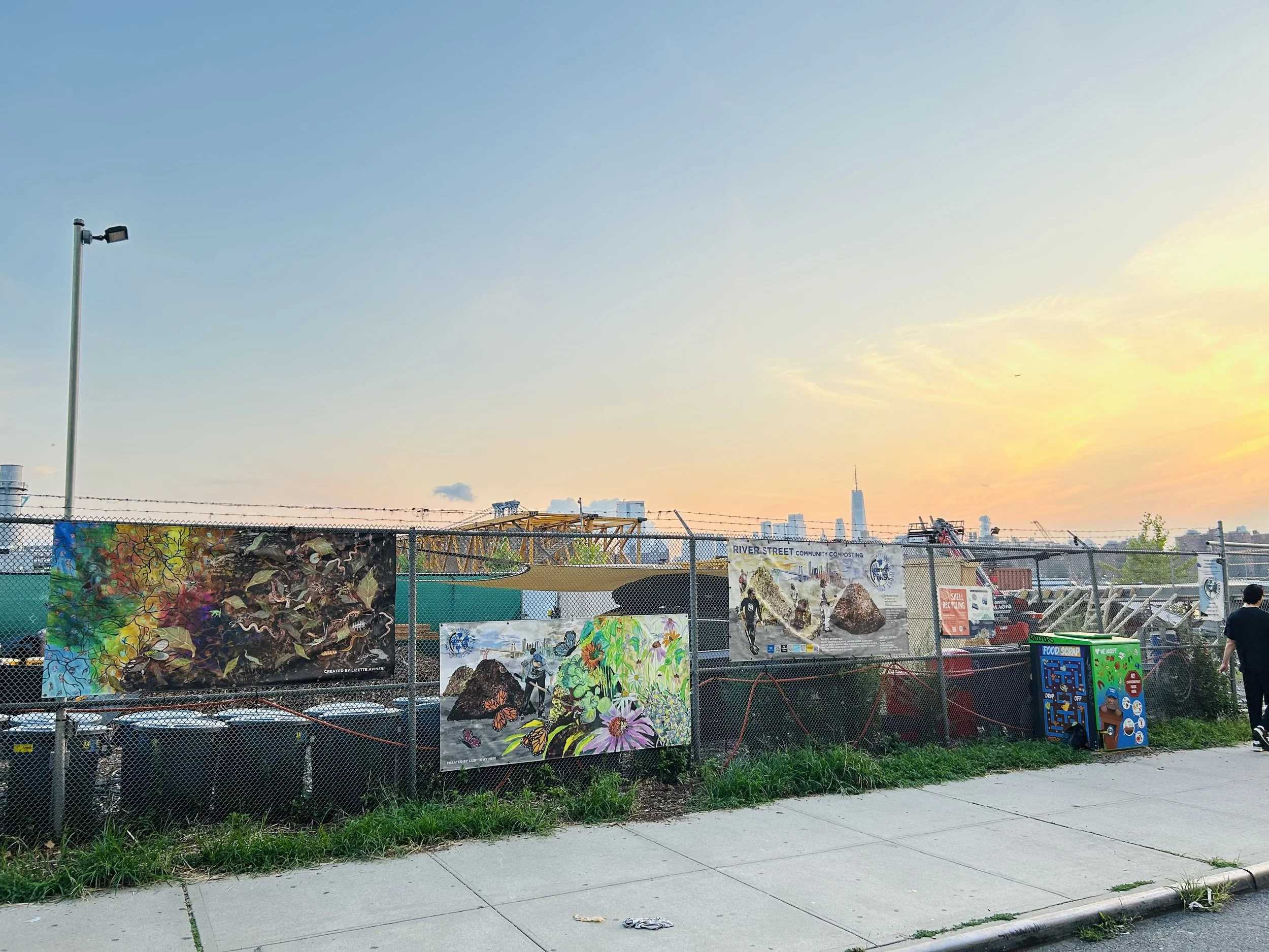 Fenced outdoor area with artwork and posters, city skyline in the background during sunset, sidewalk in the foreground.