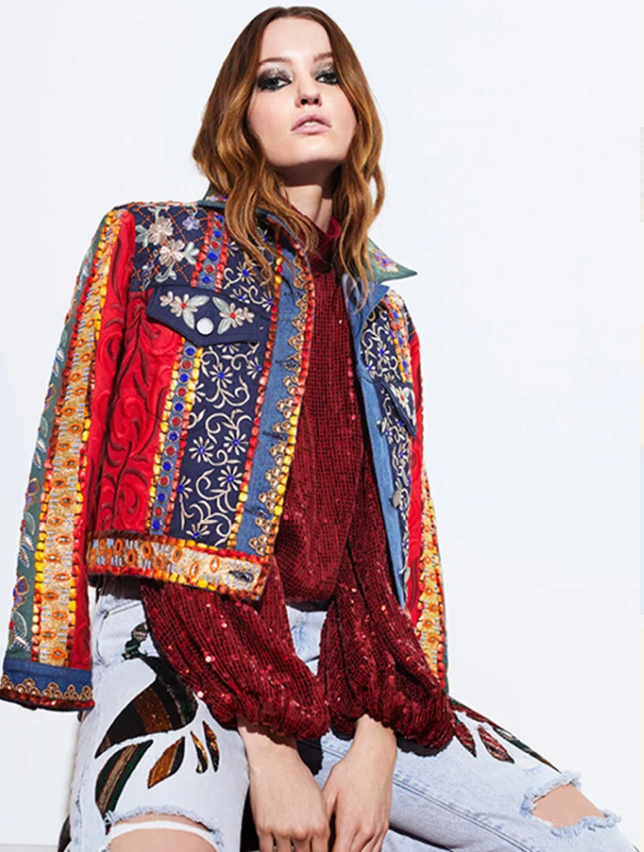 A woman with red hair wearing a colorful, embroidered jacket over a red sequined top and distressed jeans with floral embroidery.