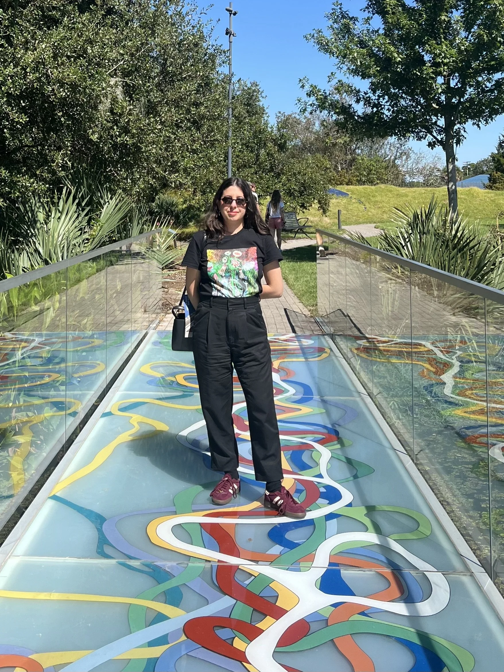 Photo of Lizette Avineri, A woman standing on a colorful, abstract walkway in a park, surrounded by greenery and trees, on a sunny day.