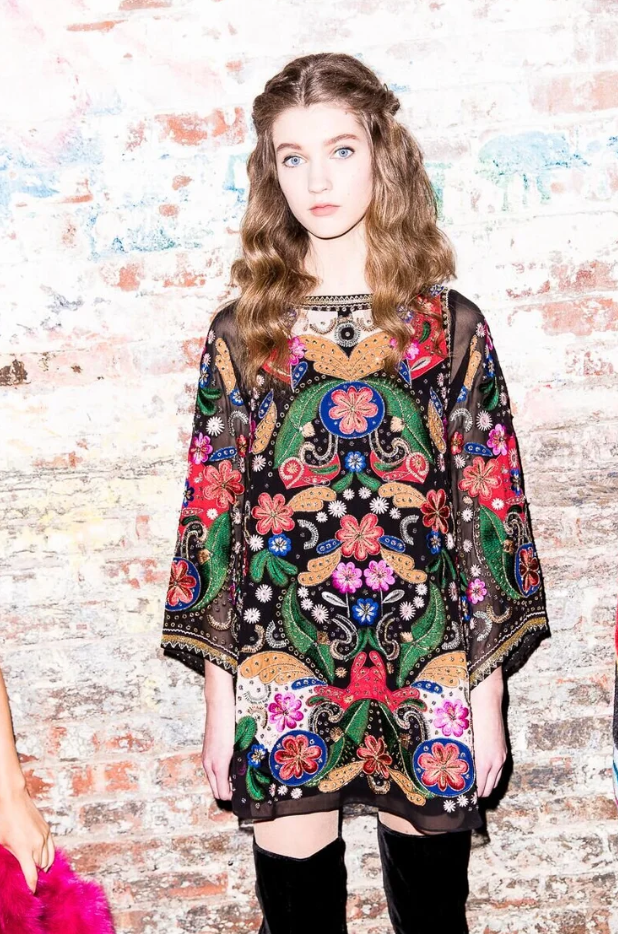 A young woman with wavy brown hair and blue eyes stands against a white and red brick wall, wearing a black embroidered dress with colorful floral and folk art designs, and black thigh-high boots.