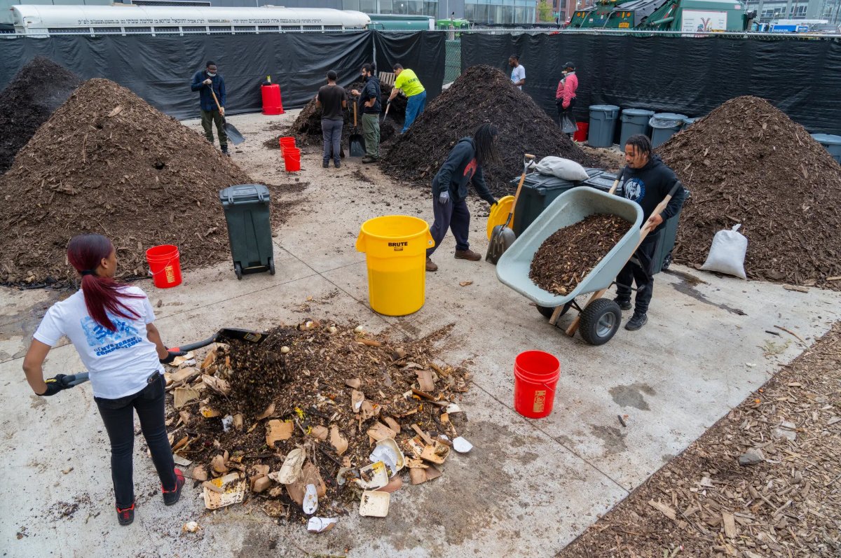 People working on a community project involving soil and mulch, with piles of dirt, shovels, wheelbarrows, and red buckets outdoors.