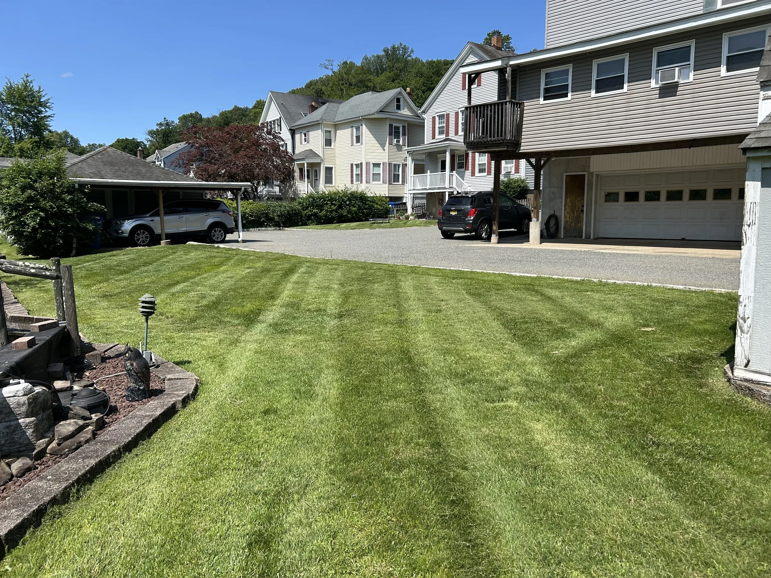 A well-maintained front yard with lush green grass and visible mowing lines, with a small garden bed on the left side, and houses in the background including a large multi-story home with a parking area and cars.