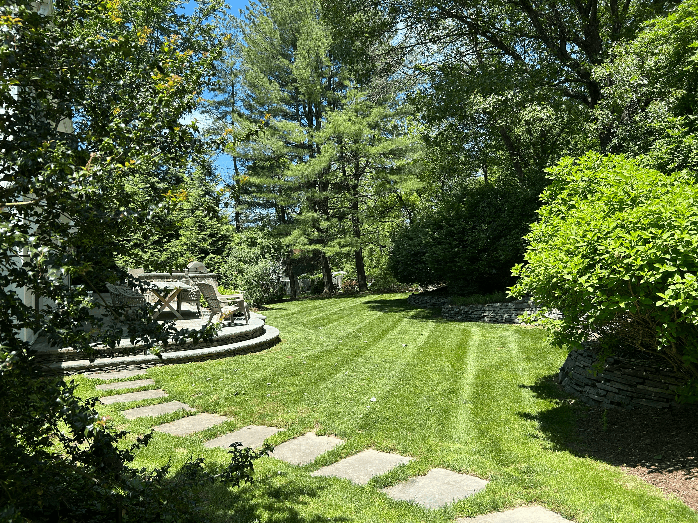 A lush green backyard with a neatly mowed lawn, stone pathway, and outdoor seating area surrounded by trees and bushes.