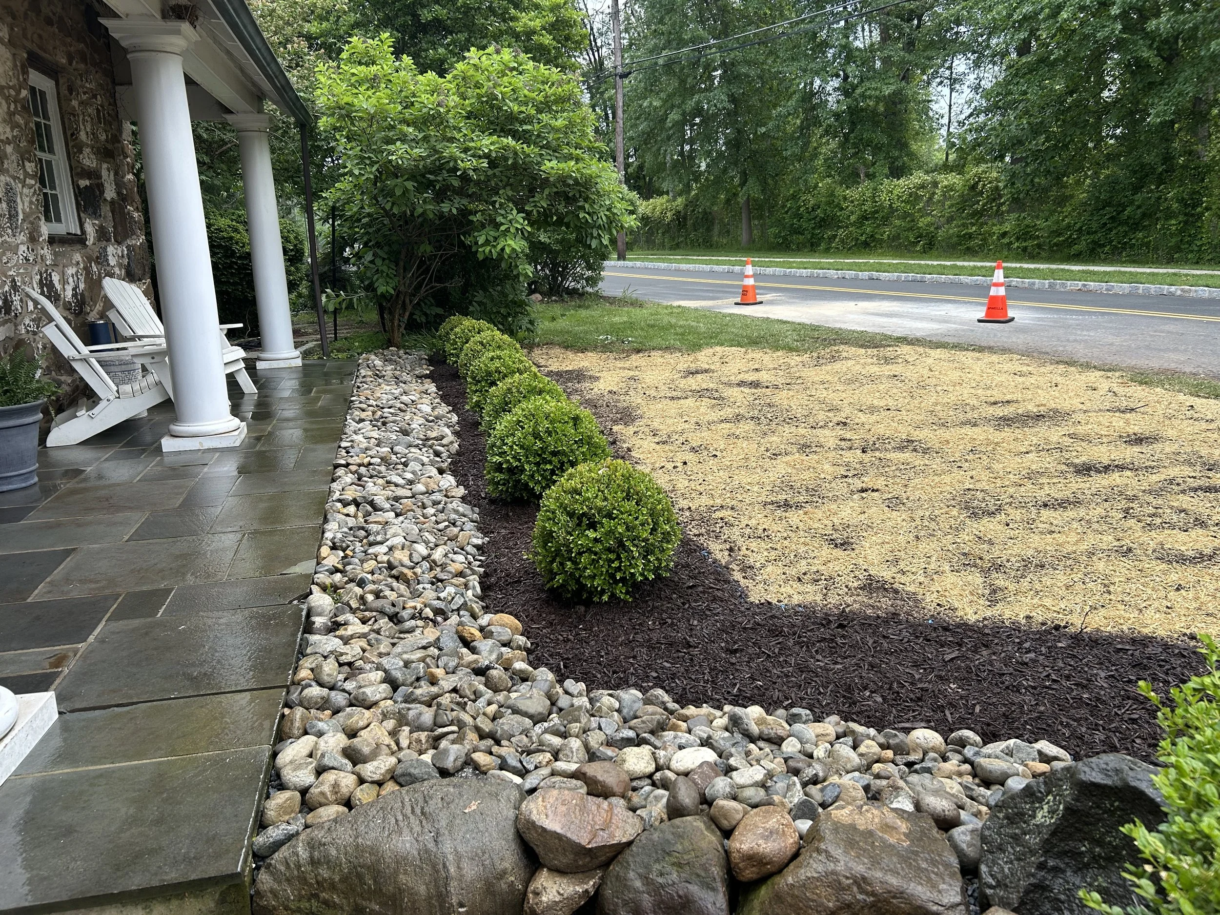 View of a front yard with a stone porch, potted plants, green shrubs, and a landscaped area with rocks and mulch. A road with traffic cones is visible in the background.