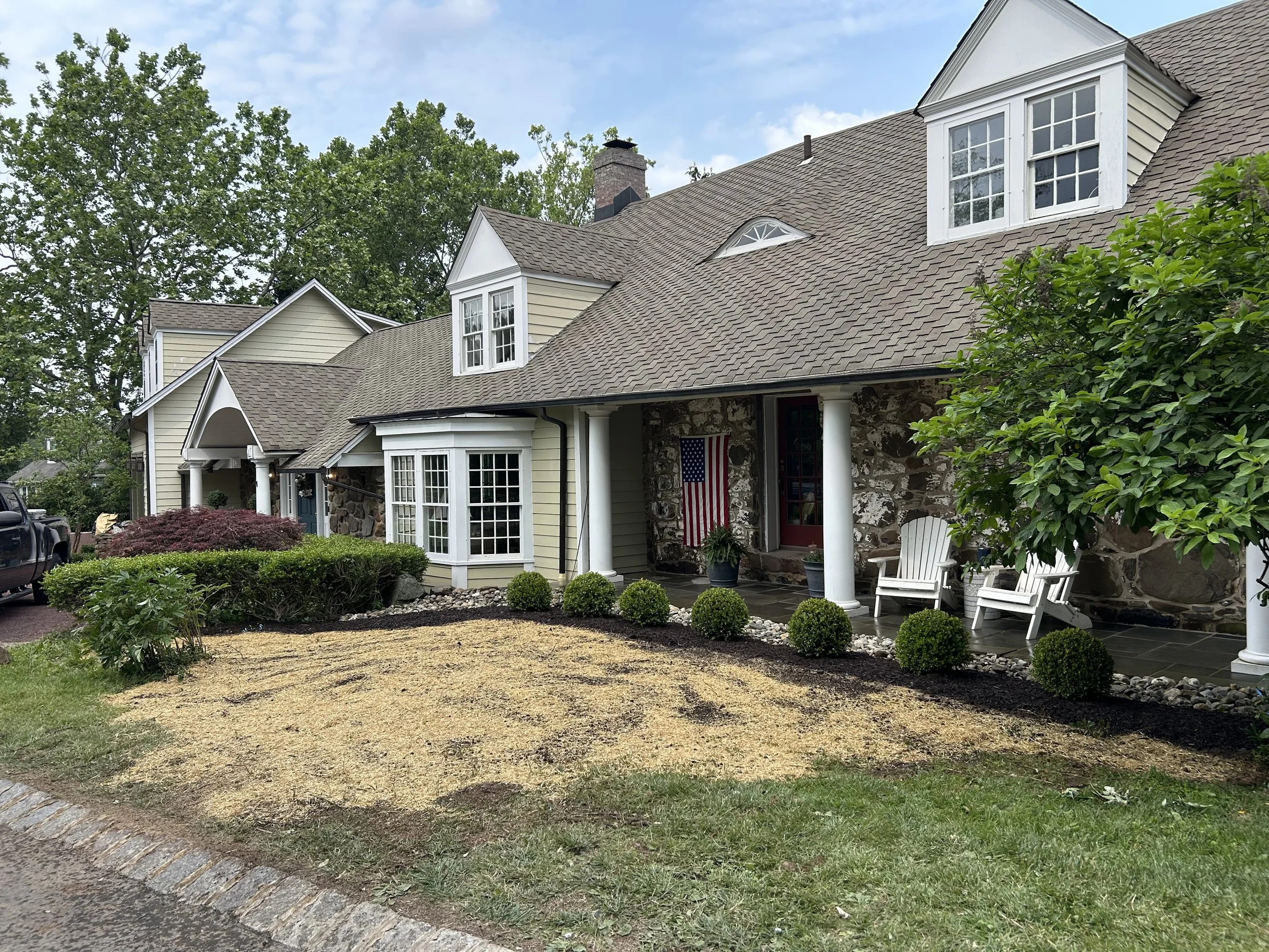 Front view of a house with light yellow siding, stone accents, a brown shingled roof, and white window trim. The porch has two white chairs, an American flag, and two potted plants. The yard has a small tree, shrubs, and a mulched bed with a layer of