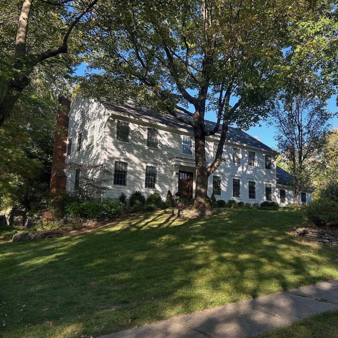 White two-story house with multiple windows, surrounded by trees and a well-maintained lawn, with shadows from the trees on the grass and house.