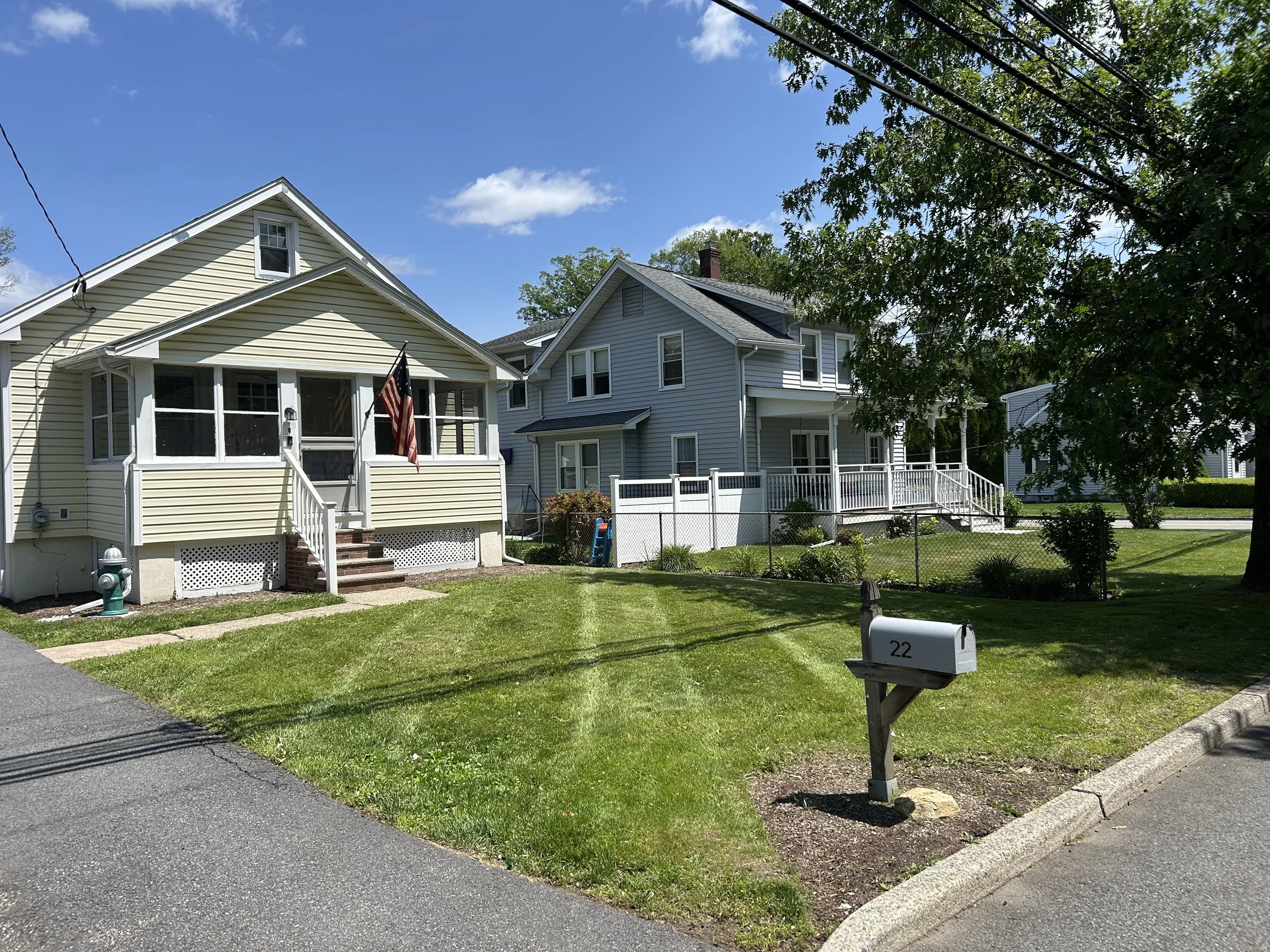 A suburban neighborhood street with two houses, one yellow and one gray, each with a small porch, surrounded by green lawns and trees, under a clear blue sky.