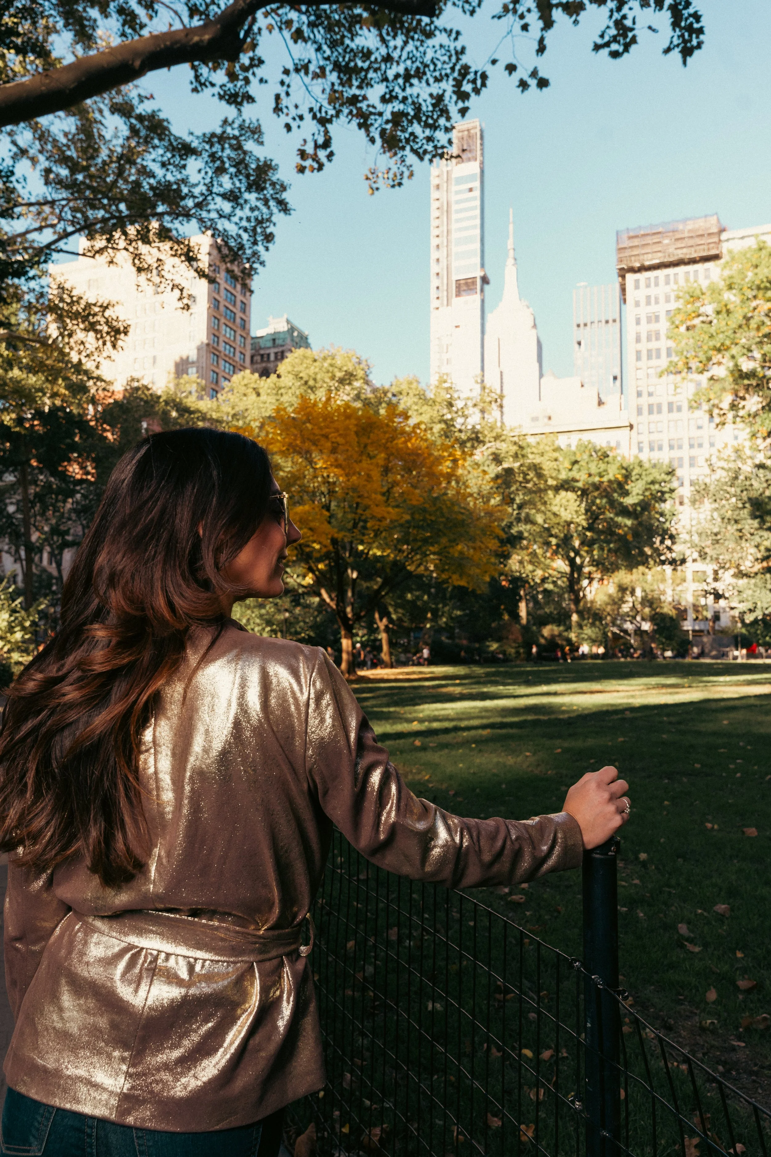 Woman with long brown hair wearing a shiny gold jacket standing by a park near the empire state building.