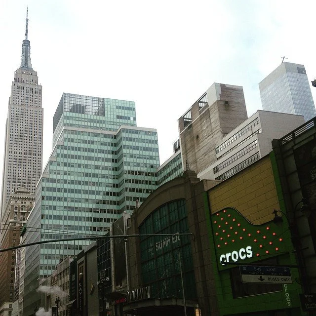 Cityscape of New York City with tall buildings, including the Empire State Building, and a Crocs store sign in the foreground.