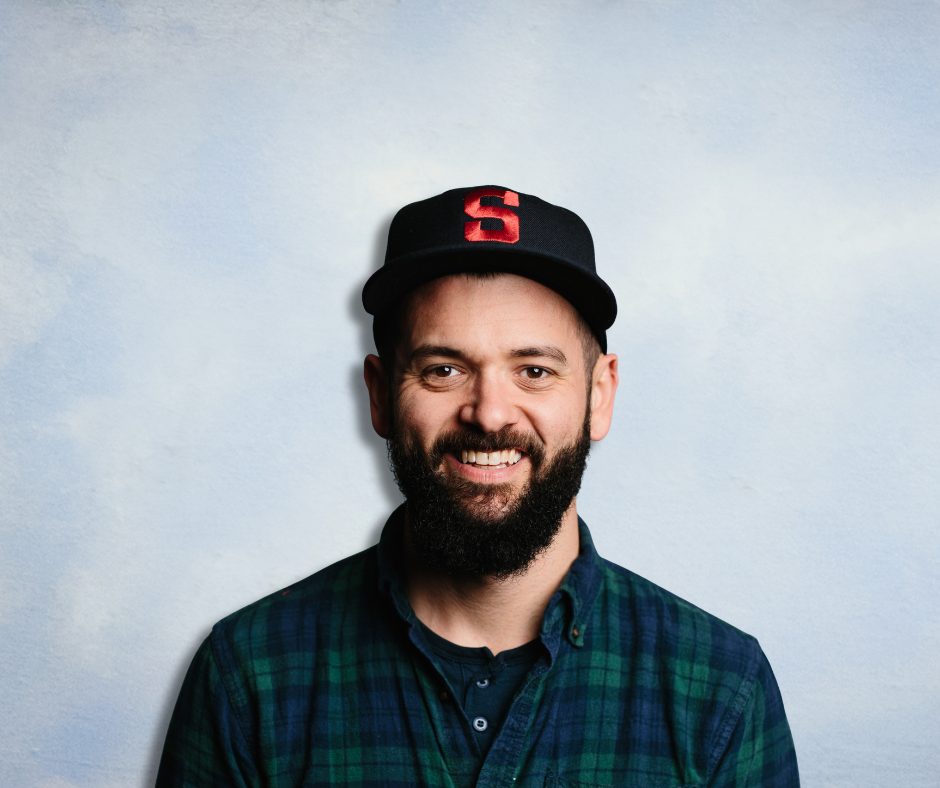 A man with a beard and mustache wearing a black baseball cap with a red letter S and a green plaid shirt, smiling at the camera against a light blue background.