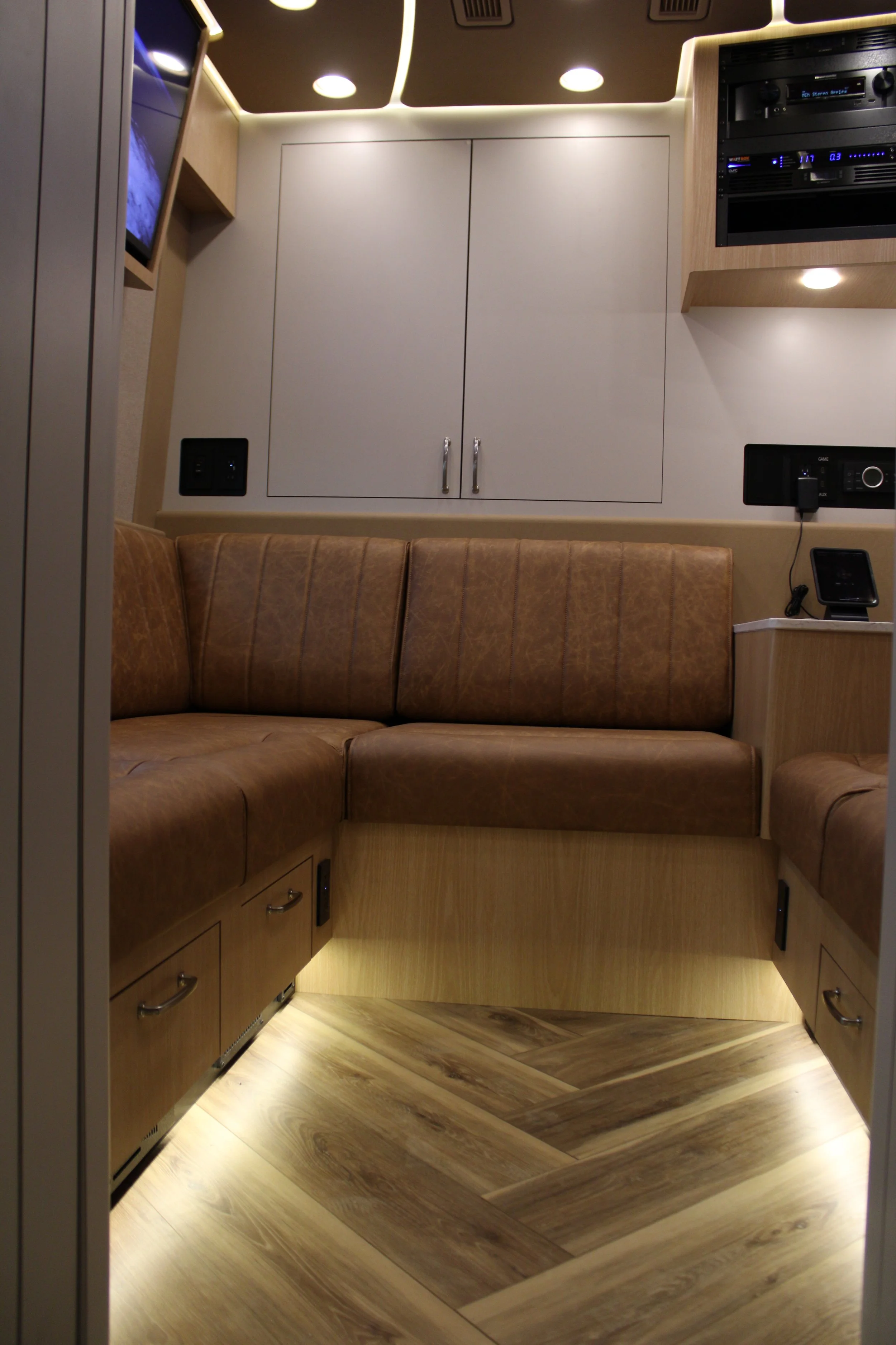 Interior of a lounge or small room with beige walls, a curved brown leather sofa, wooden flooring, ceiling lights, a cabinet, and electronic equipment.