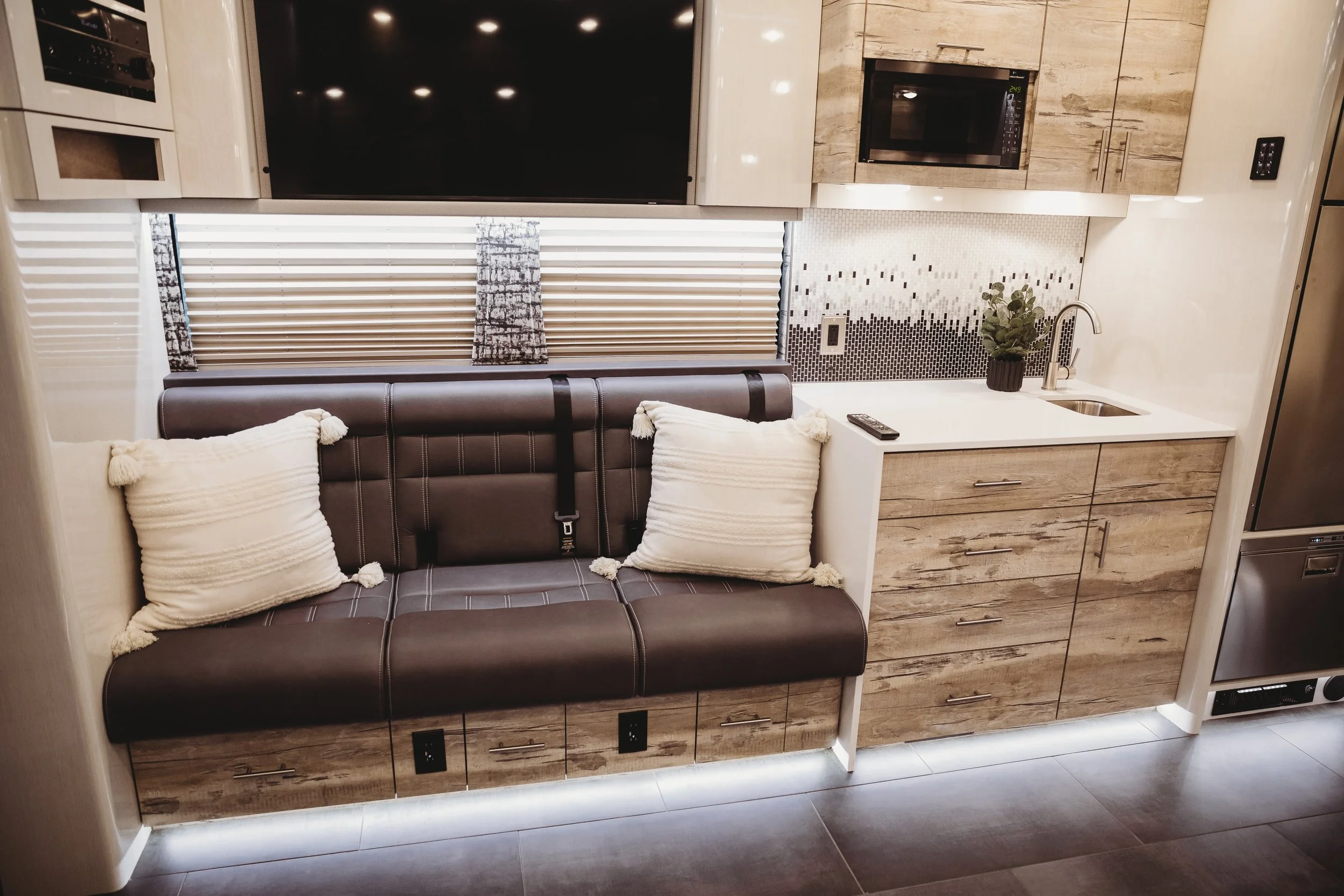 Interior of a modern compact kitchen with a dark leather sofa, beige wooden cabinet, small white countertop with a sink, potted plant, remote control, and a wall-mounted microwave. Window with blinds behind the sofa.