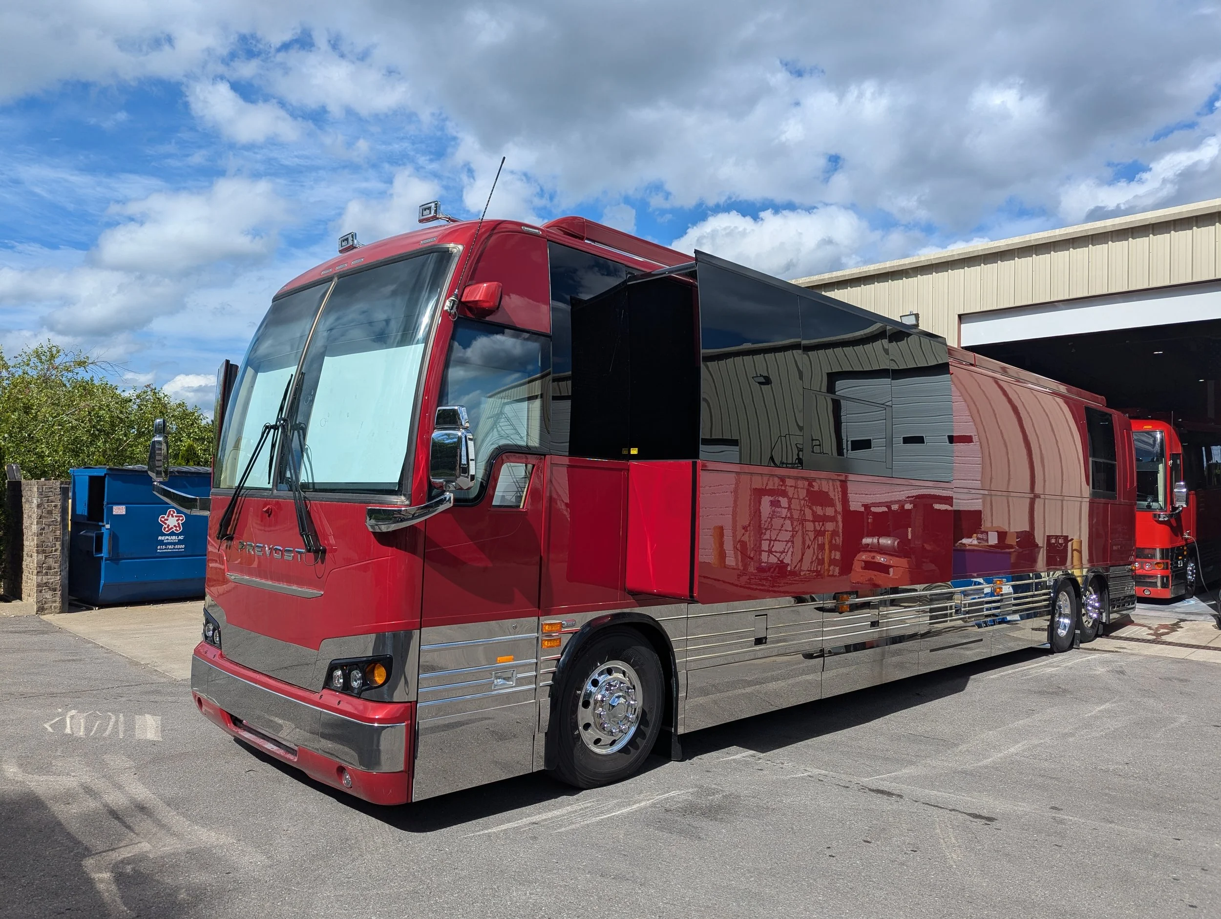 A large red and black luxury RV parked outside a service building with a partly cloudy sky in the background.