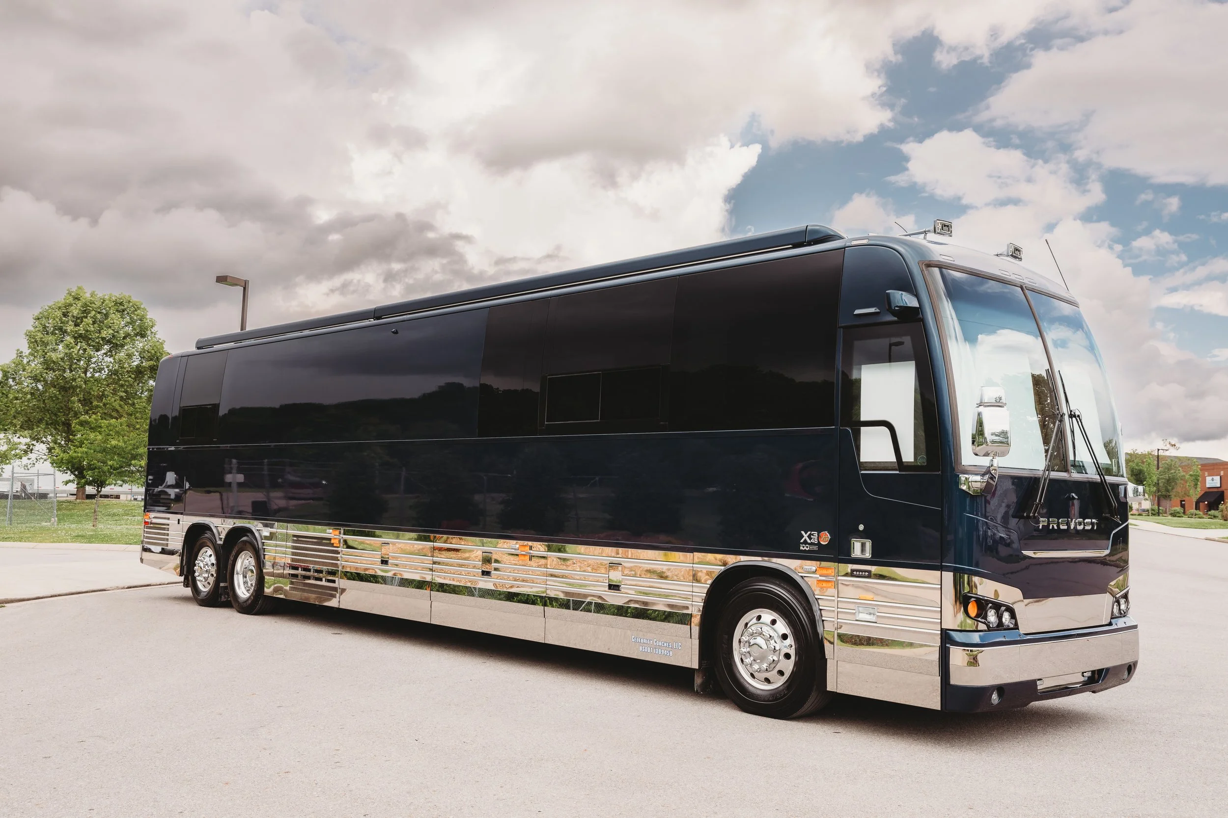 Black luxury tour bus parked in an open lot with cloudy sky and trees in the background.