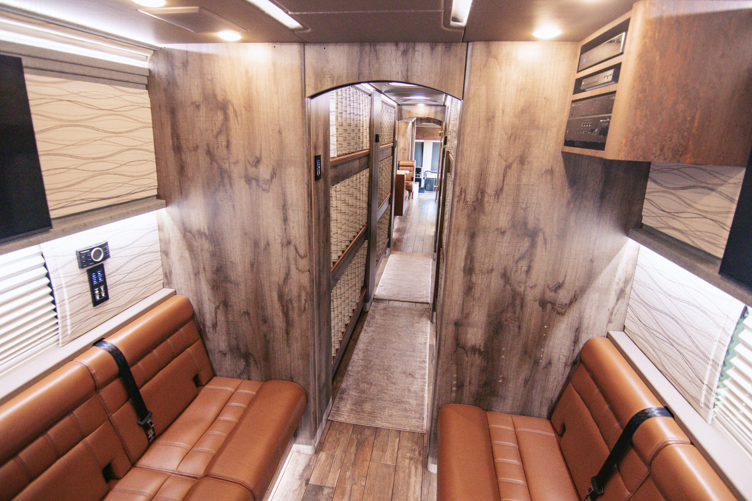 Interior view of a compact camper or recreational vehicle with two brown leather bench seats facing each other, wood-paneled walls, and a narrow walkway leading to the living area with a small table, a window with blinds on each side, and built-in ca