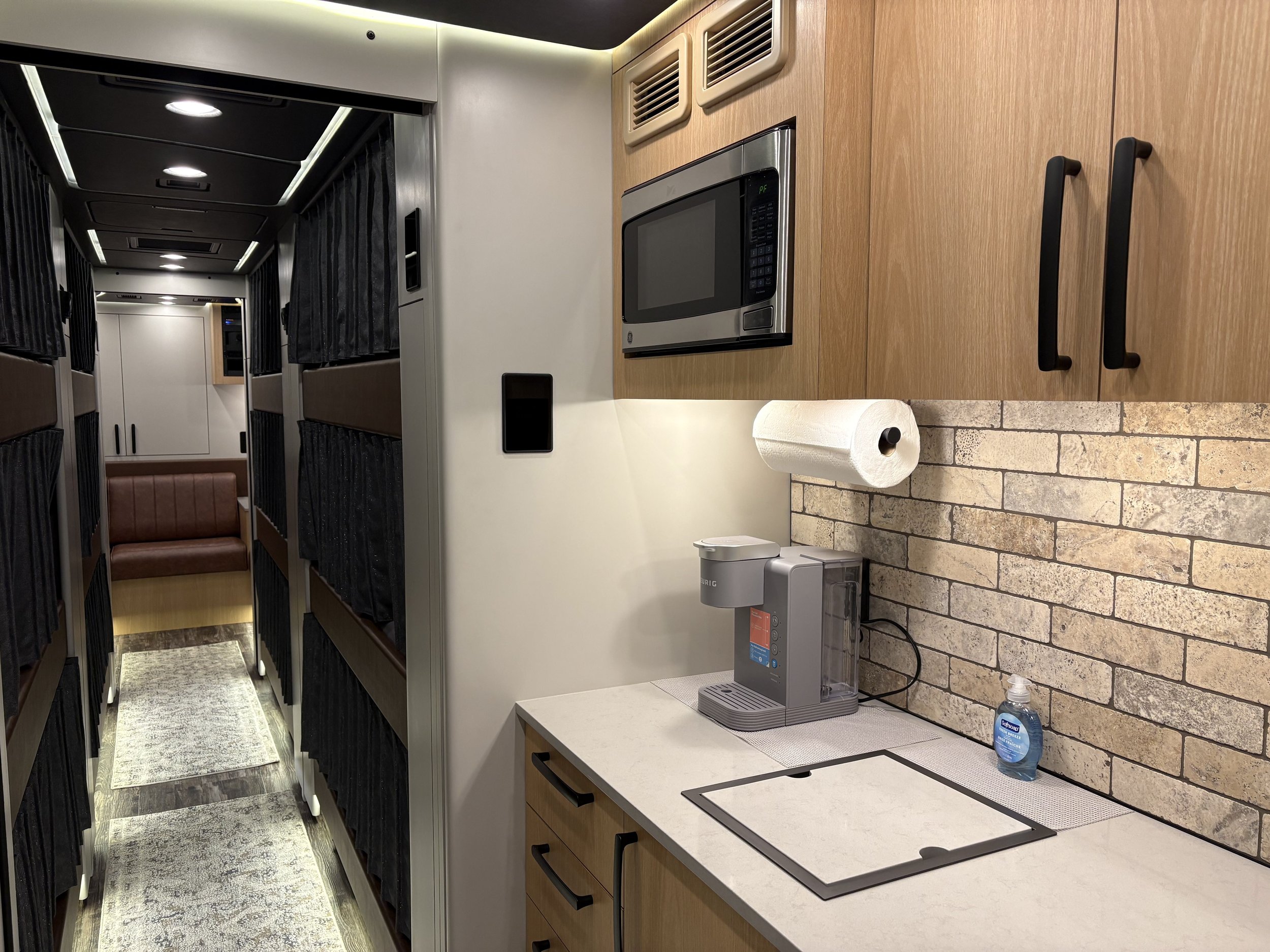 Cozy interior of a capsule hotel with sleeping pods on the left and a small kitchen area on the right, featuring a microwave, paper towel roll, water dispenser, and hand sanitizer.