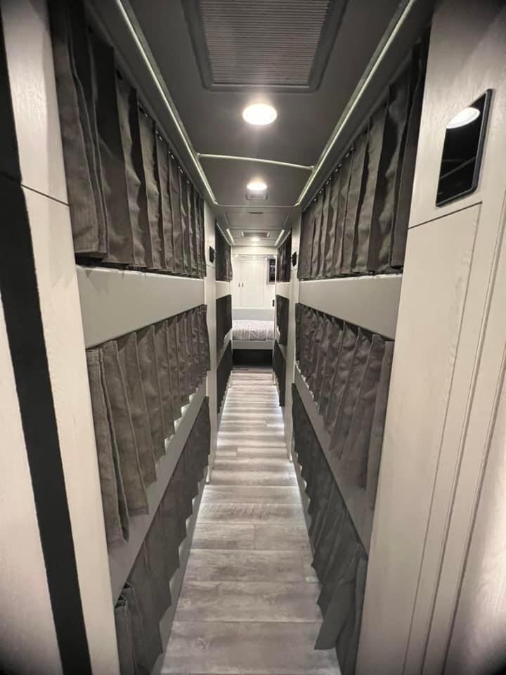 Narrow hallway inside a mobile home or RV with multiple small storage compartments with curtains on either side and a bed at the end.