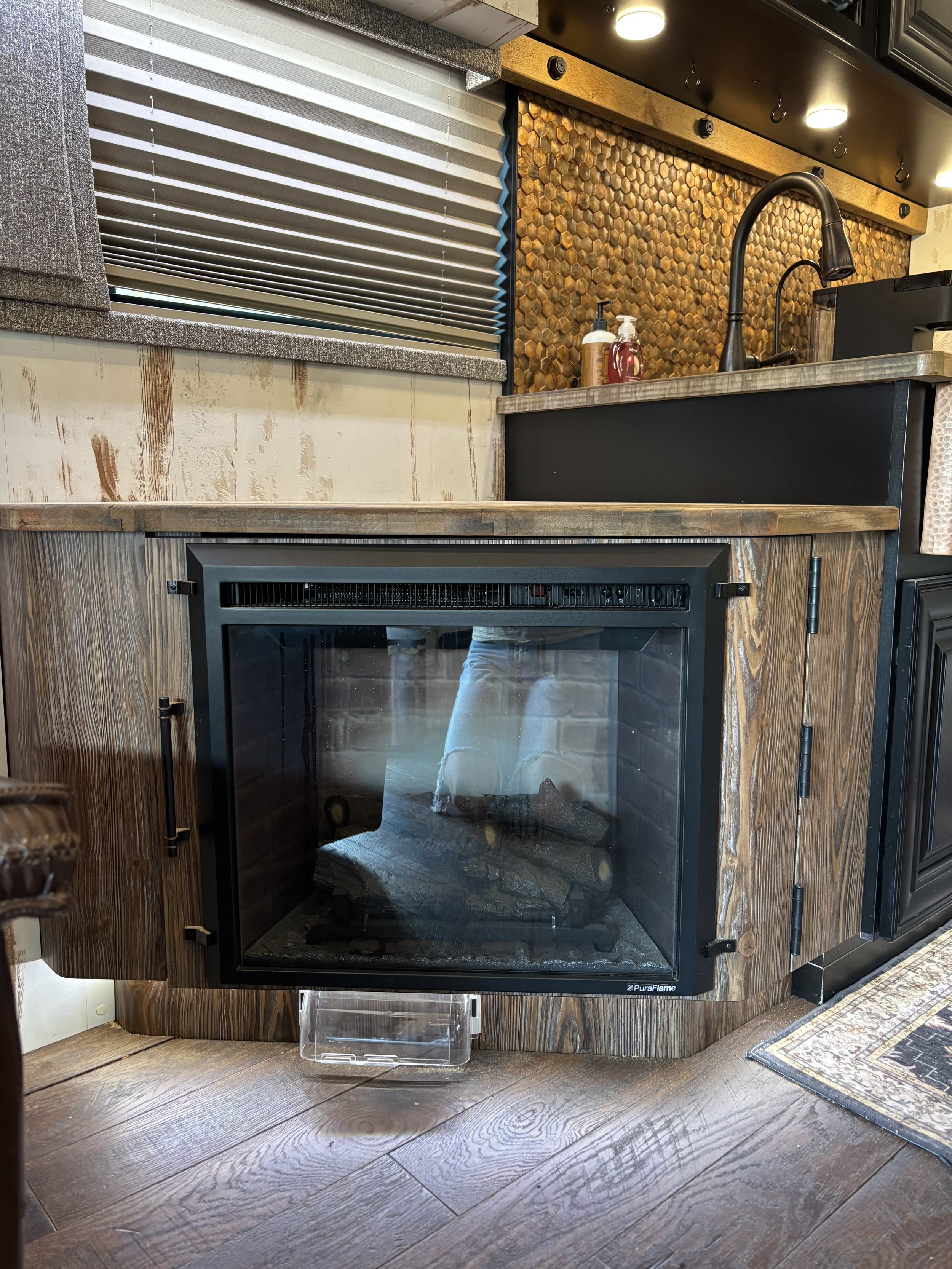 A cozy kitchen corner featuring a wood-burning fireplace with a glass door, surrounded by wooden cabinetry and a countertop with a sink. There is a window with blinds above the countertop and bottles of soap near the sink. The floor is wooden, and th