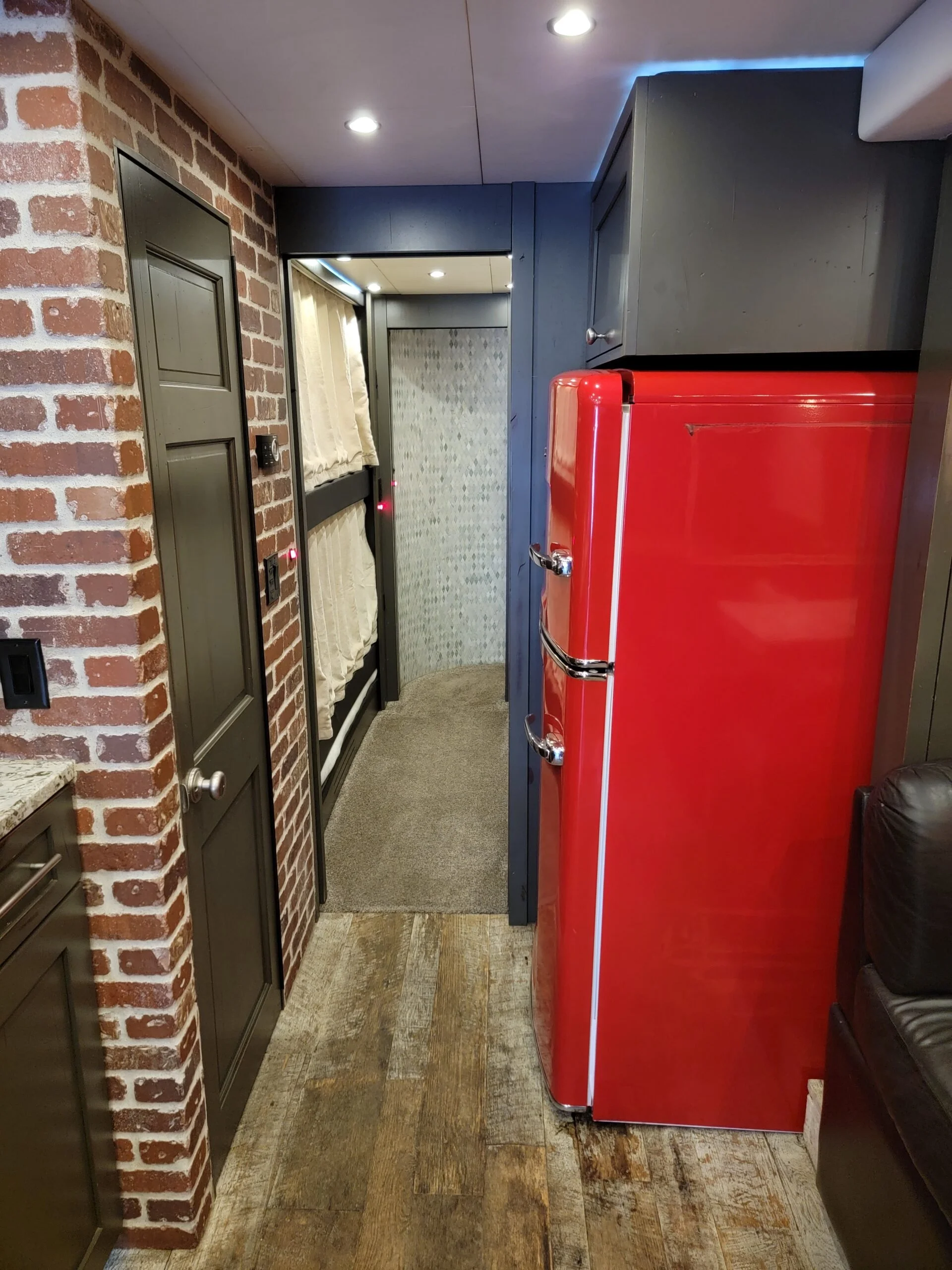A vintage red refrigerator in a kitchen with brick and wooden flooring, dark cabinets, a doorway with curtains, and ceiling lights.