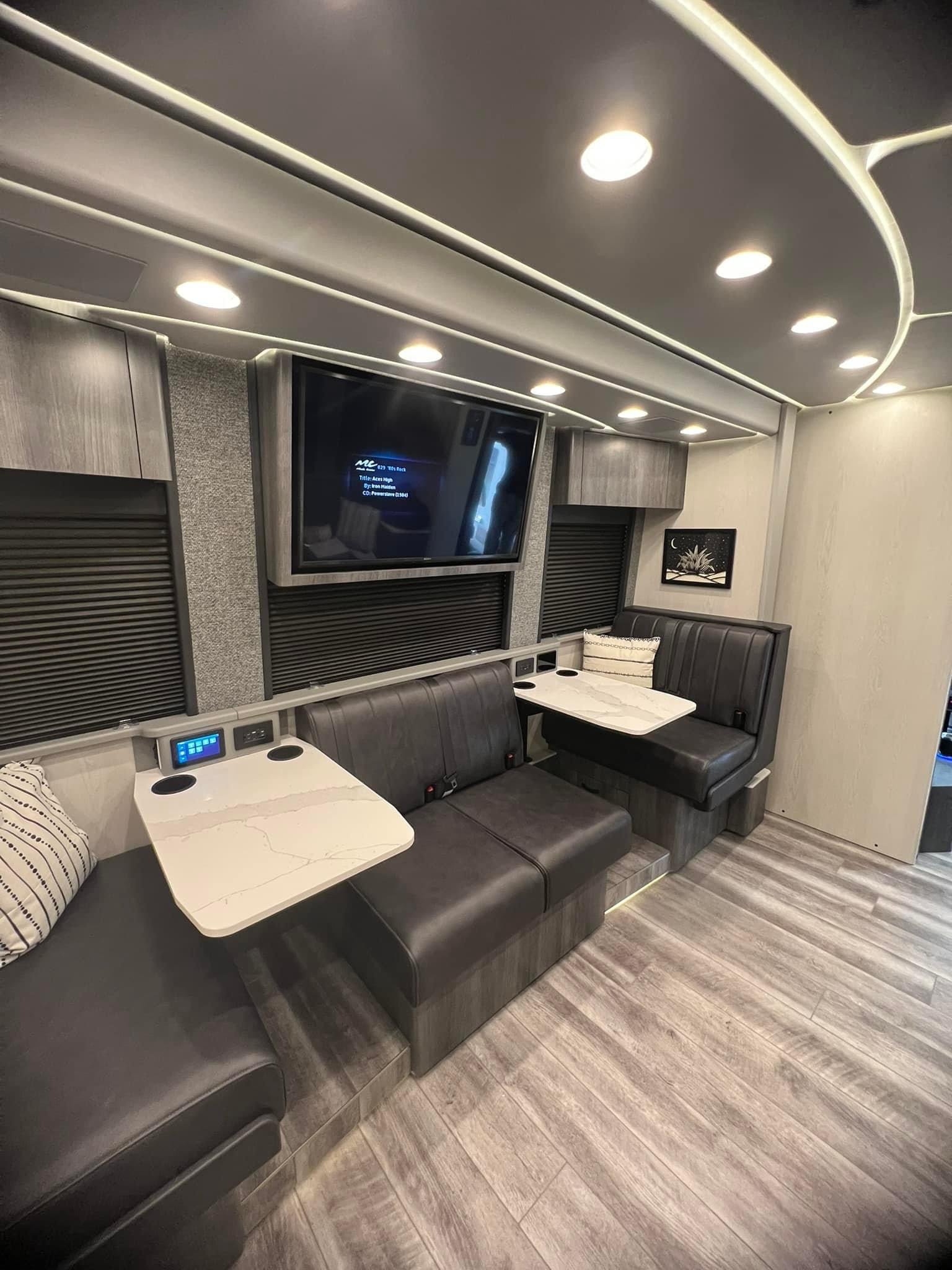 Interior of a modern L-shaped lounge with black cushioned seating, two small marble-top tables, built-in lighting, a large wall-mounted television, and decorative wall art.