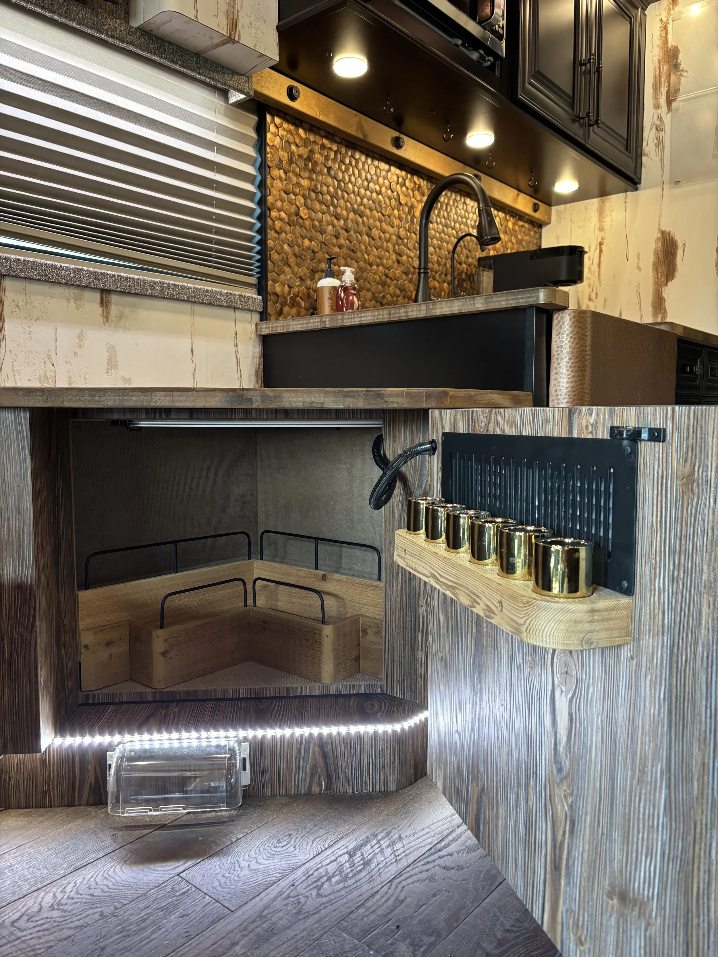 Close-up of a kitchen counter with a black sink, soap dispenser, and copper tile backsplash. Beneath the counter is an empty wooden storage area with black metal handles. To the right is a wooden organizer with gold hooks, and the floor is wood.