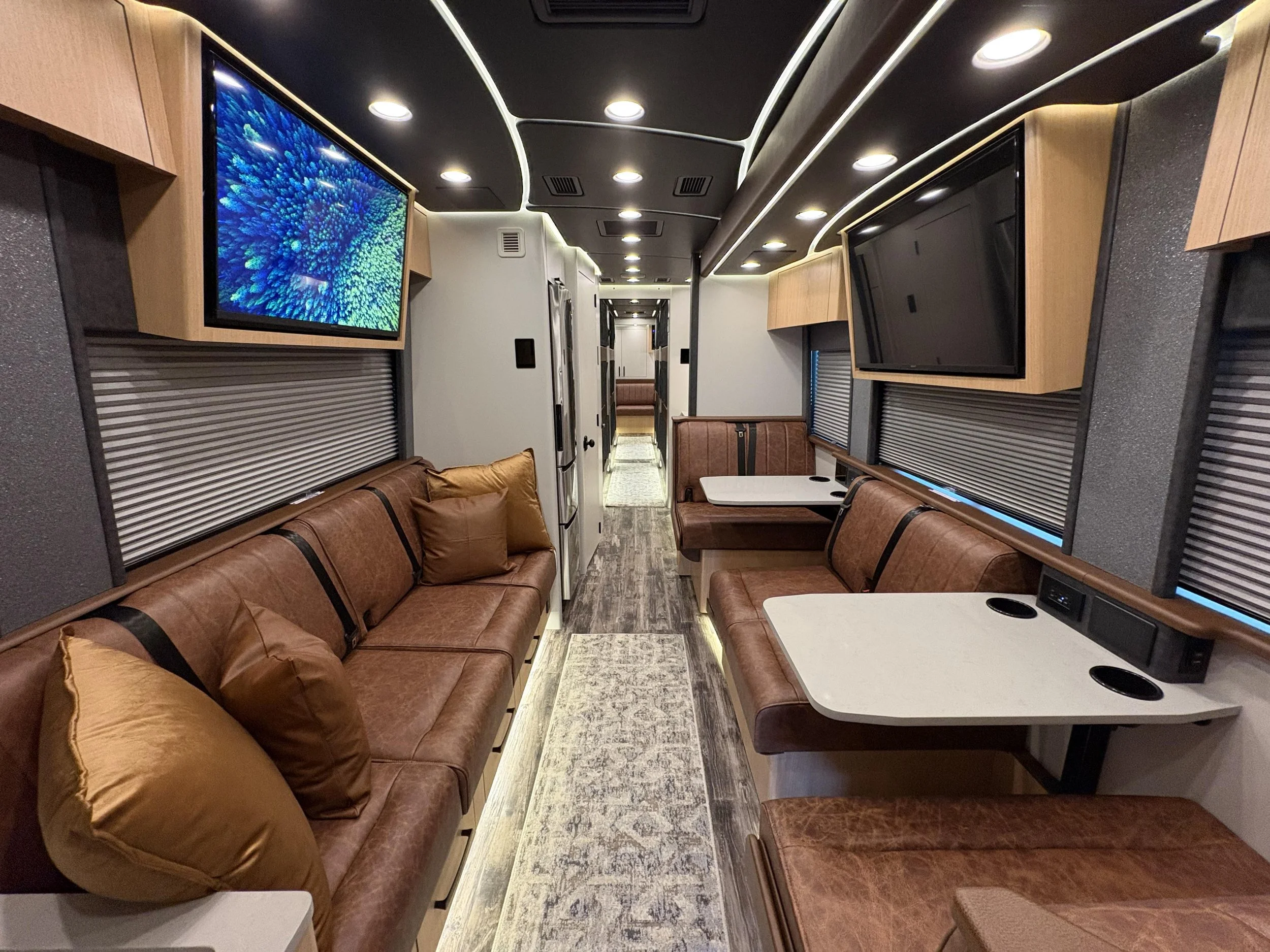 Interior of a modern recreational vehicle with leather sofas, mounted flat-screen TVs, wood cabinets, and windows with blinds.