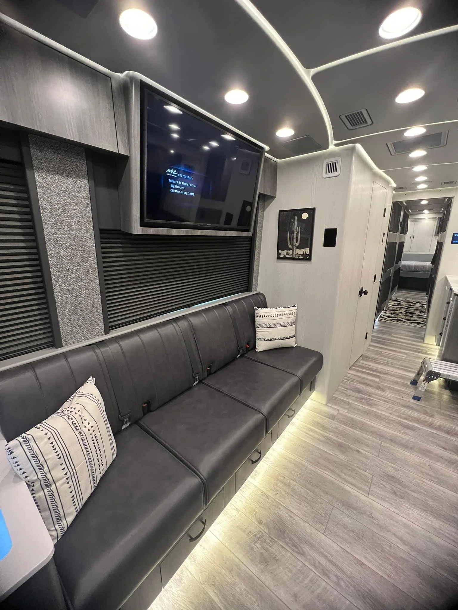 Interior of a modern RV or tiny home with a black leather sofa against a wall, decorative pillows, a wall-mounted flat-screen TV, and light-colored wood flooring. Recessed ceiling lights illuminate the space.