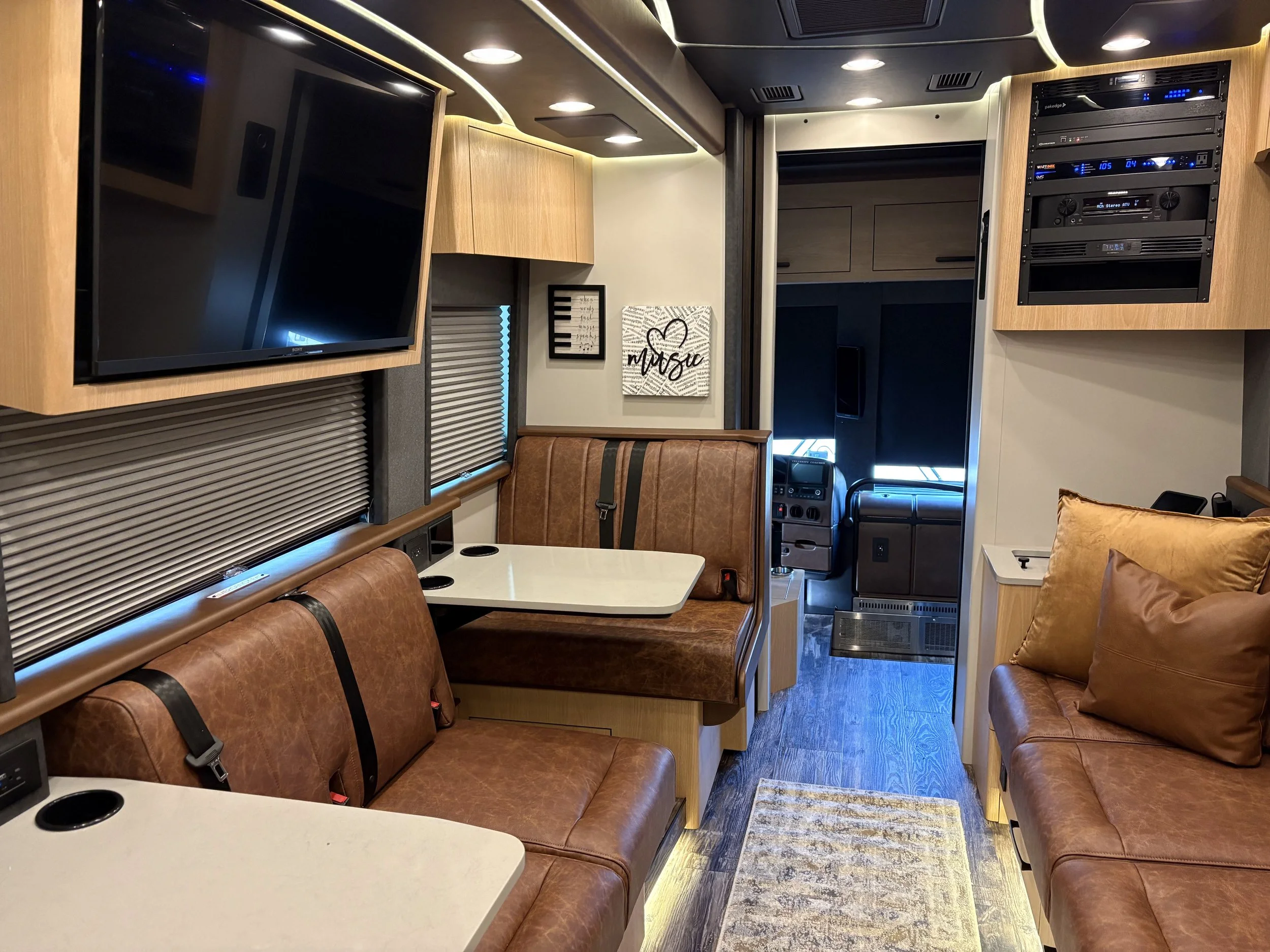 Interior of a recreational vehicle or camper with leather seating, a wall-mounted television, window blinds, and a small dining area, with a visible control panel and dashboard in the driver's cab area.