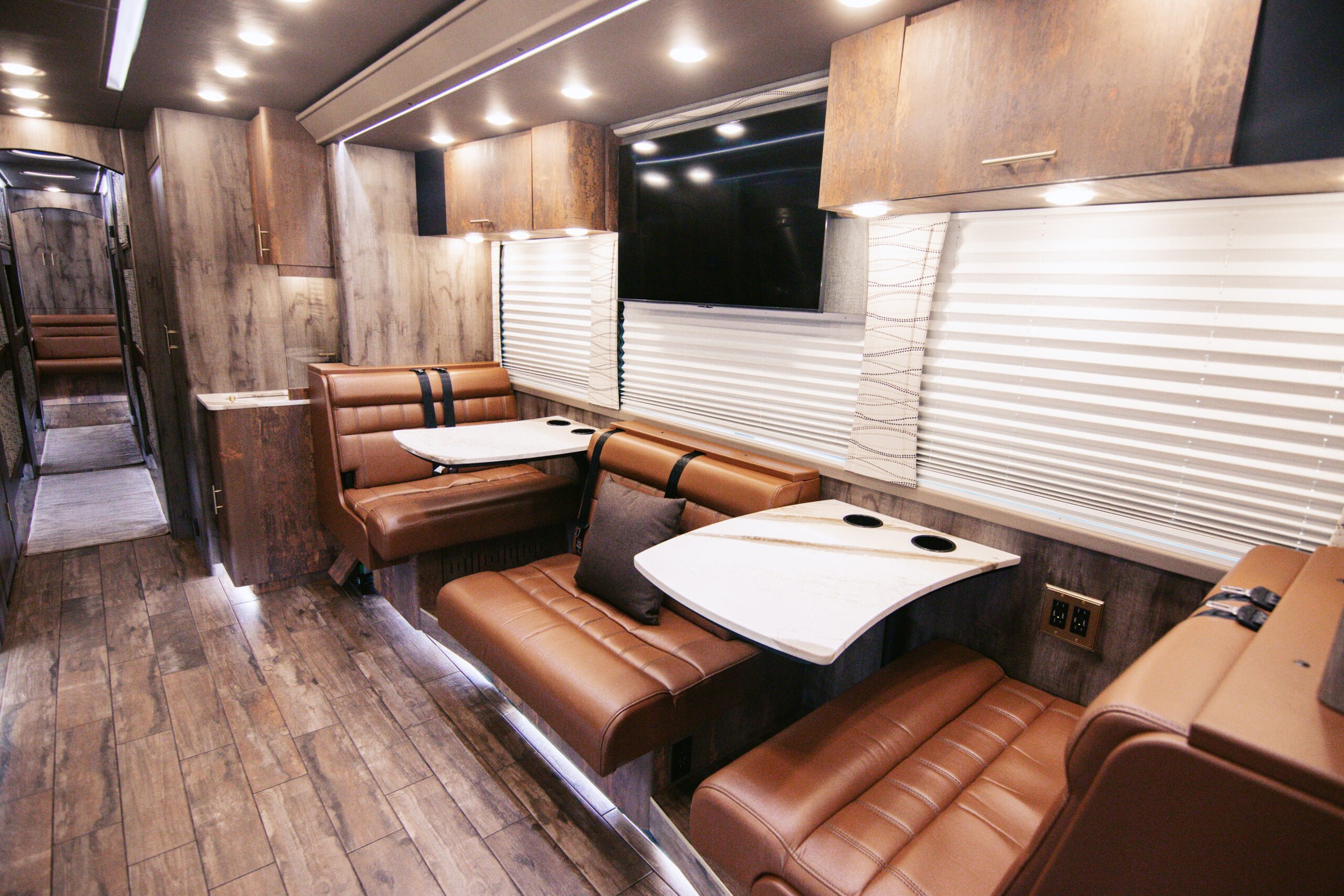 Interior of an RV or mobile home with brown leather seating, white marble tables, wood-paneled walls, and large windows with blinds.