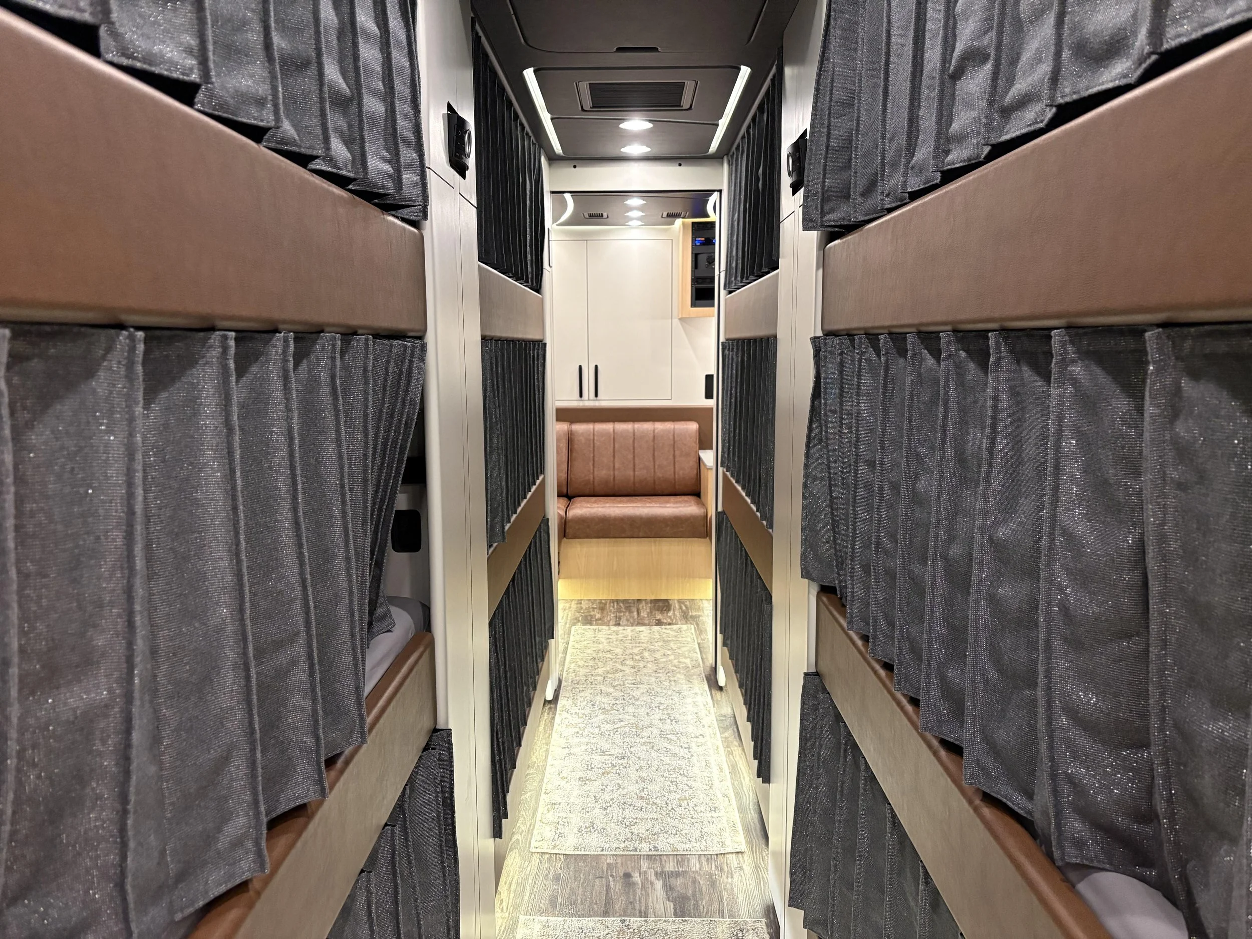 Interior of a bus or train with sleeping compartments on both sides, each with brown beds and black curtains. A small corridor leads to a seating area with a pink cushioned bench in the background.