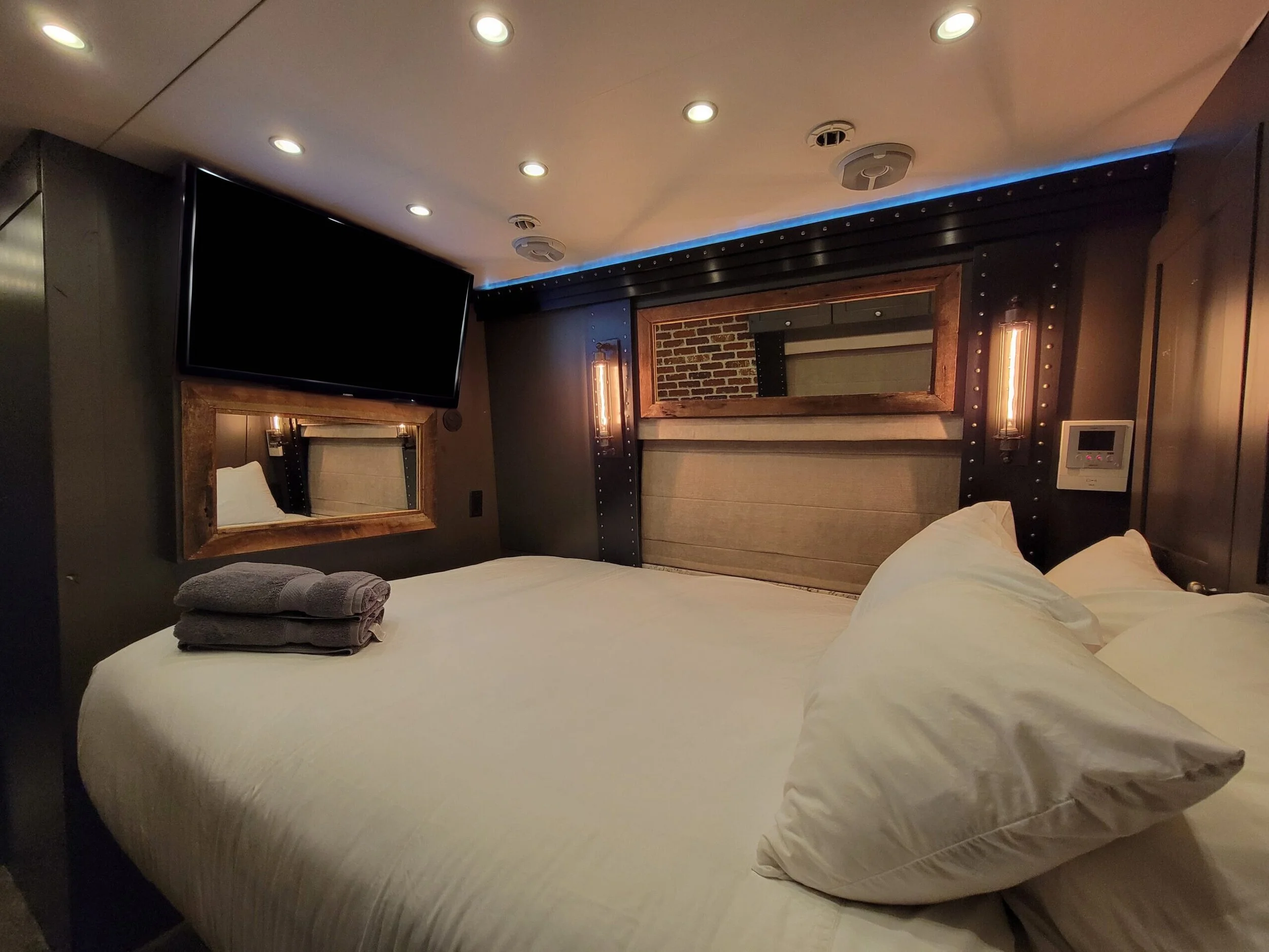 Modern hotel room with a bed, gray towels, wall mirror, large TV, illuminated wall sconces, and decorative elements.