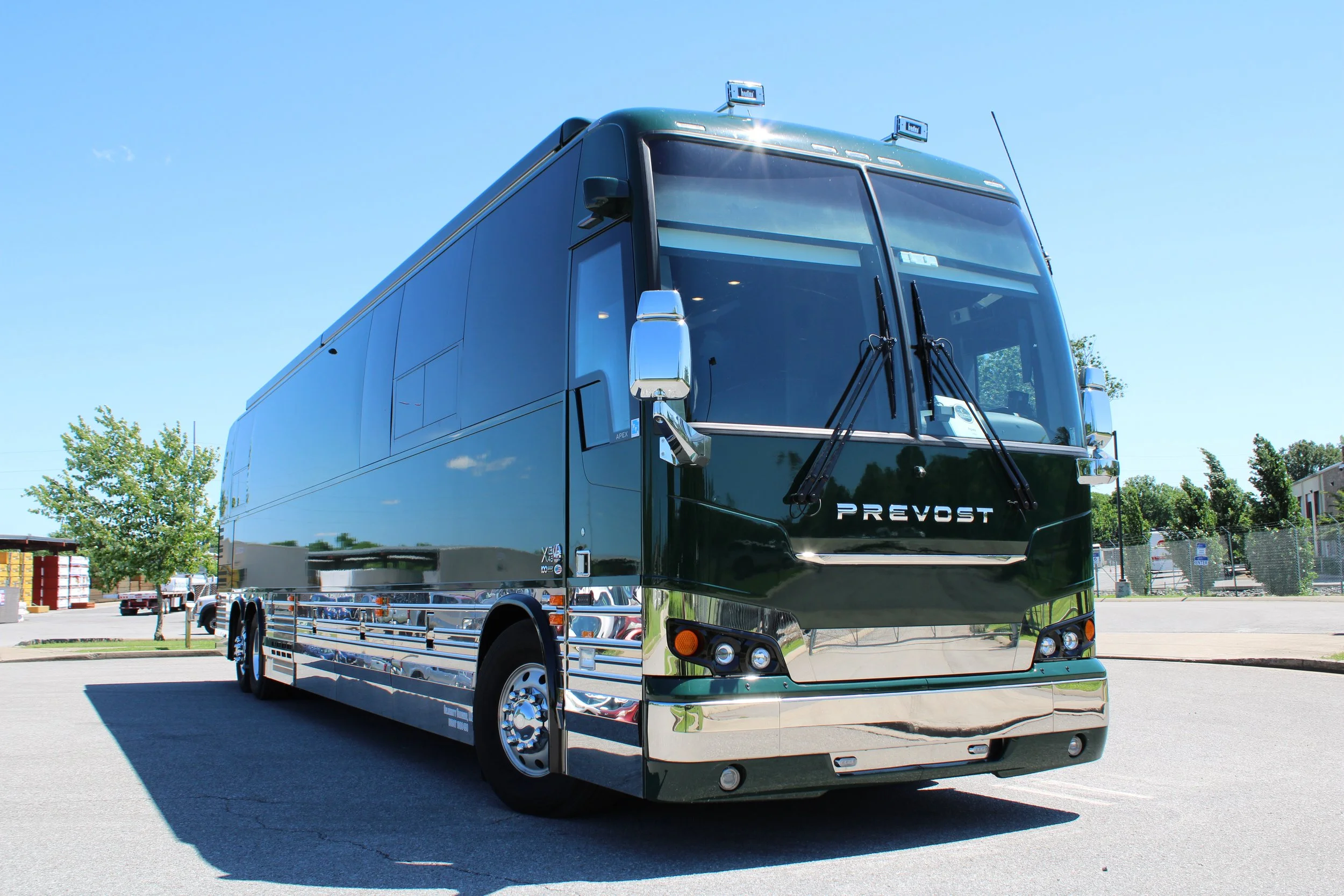 A black Prevost bus parked in a lot on a sunny day, with trees and a fence in the background.