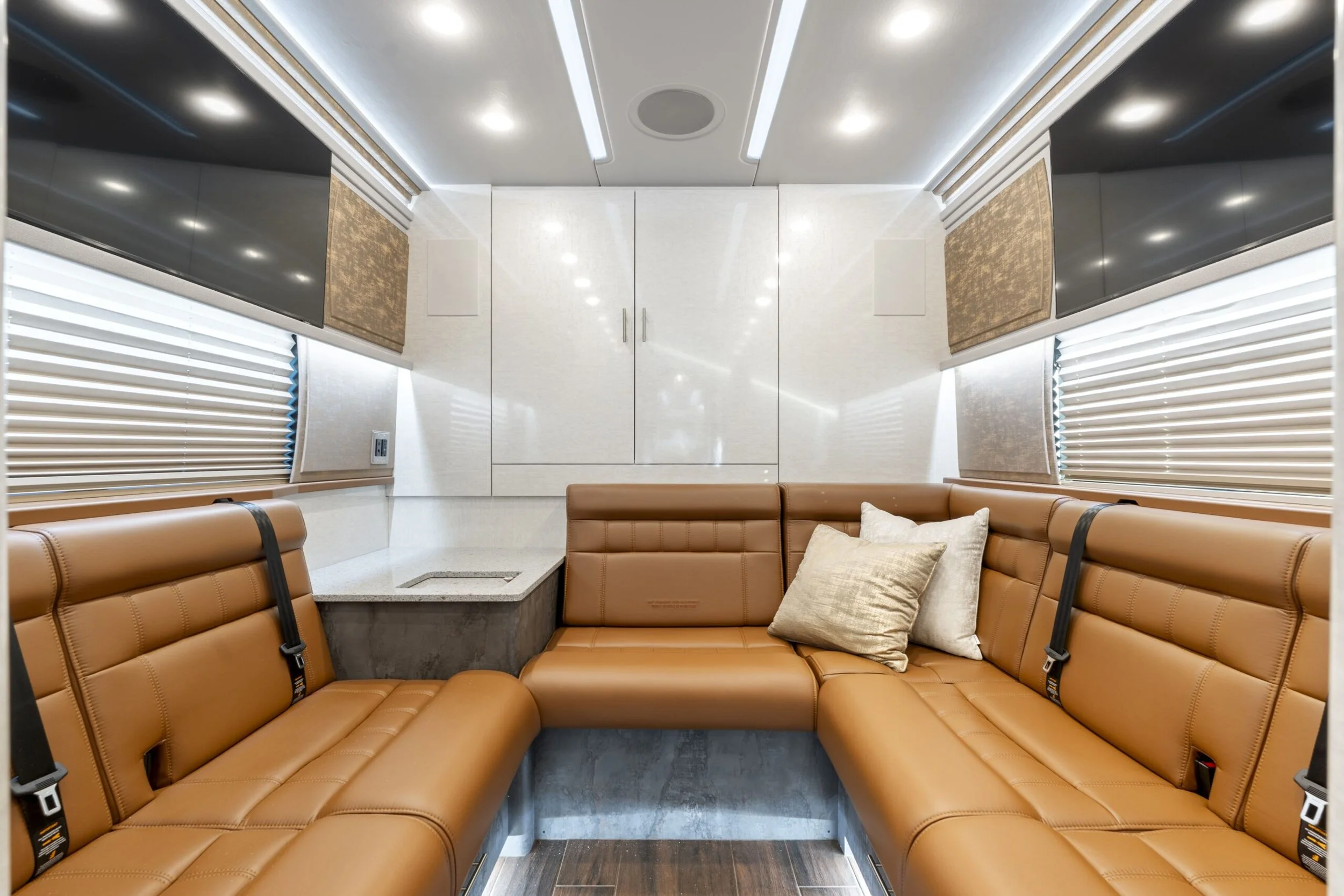 Interior of a modern bus or camper with tan leather seating, pillows, window blinds, and overhead cabinets.