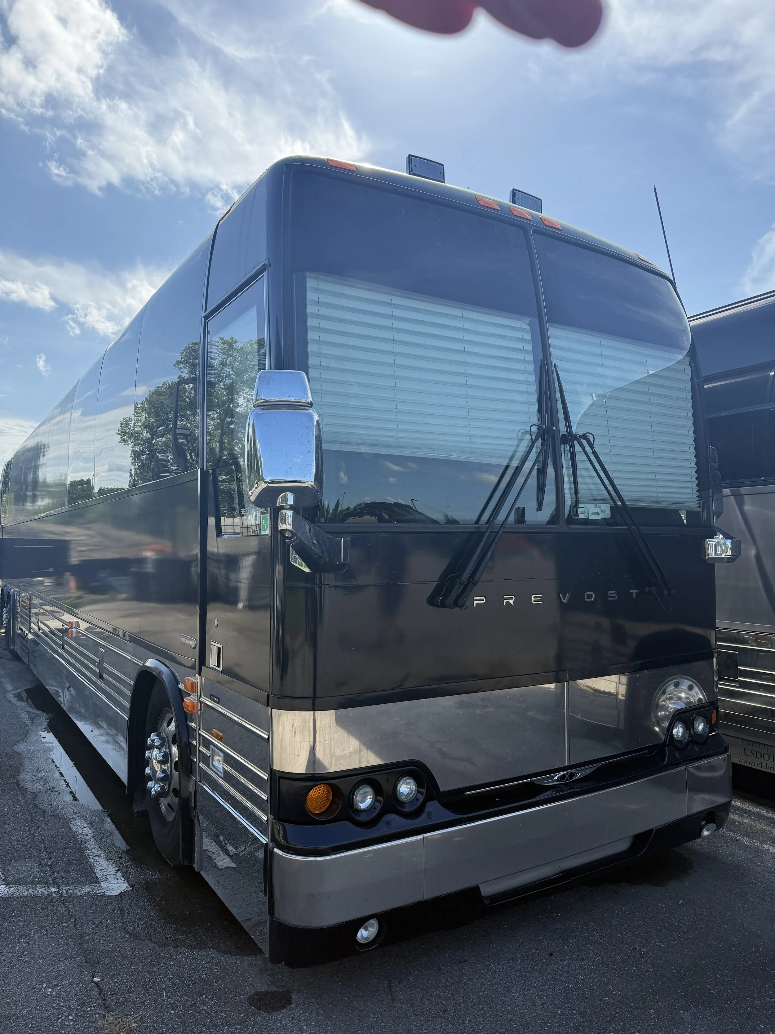 Black luxury bus parked on the street under a blue sky with some clouds, reflecting nearby trees on its shiny surface.