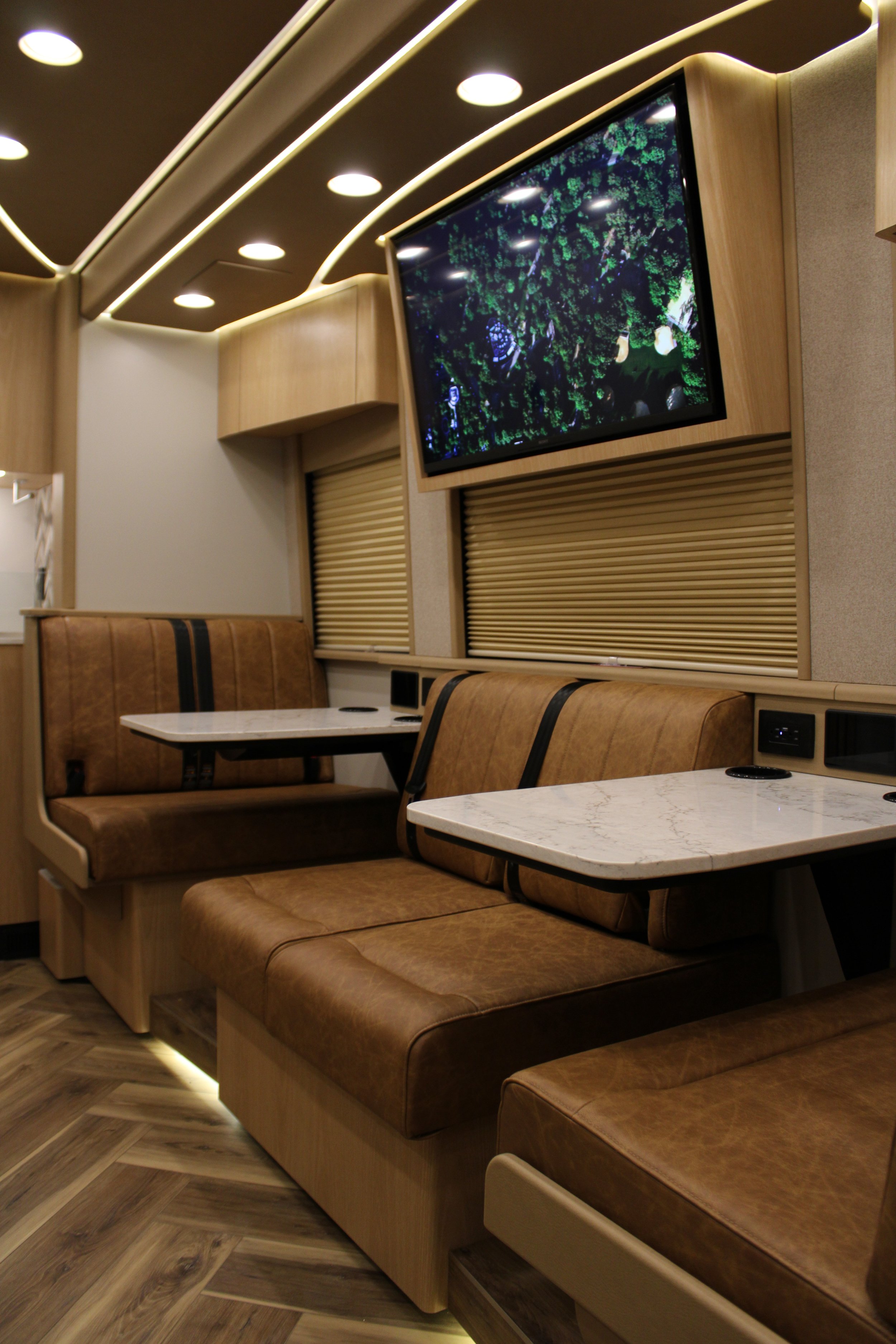 Interior of a modern recreational vehicle with leather booths, marble tables, and a wall-mounted flat-screen TV showing greenery.