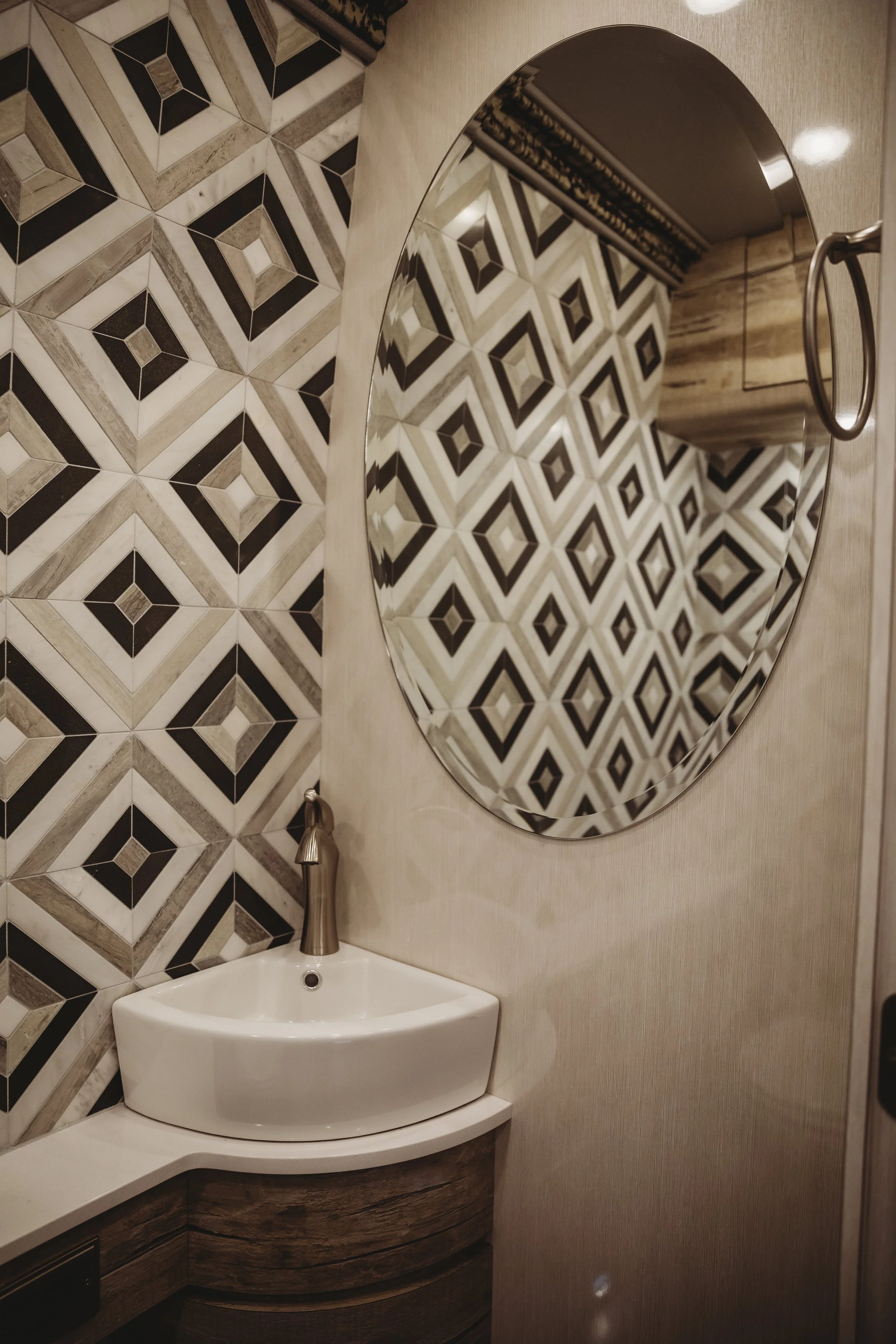 A small bathroom vanity with a white sink, a curved wooden cabinet, a large round mirror, and dark geometric patterned tiles on the wall.