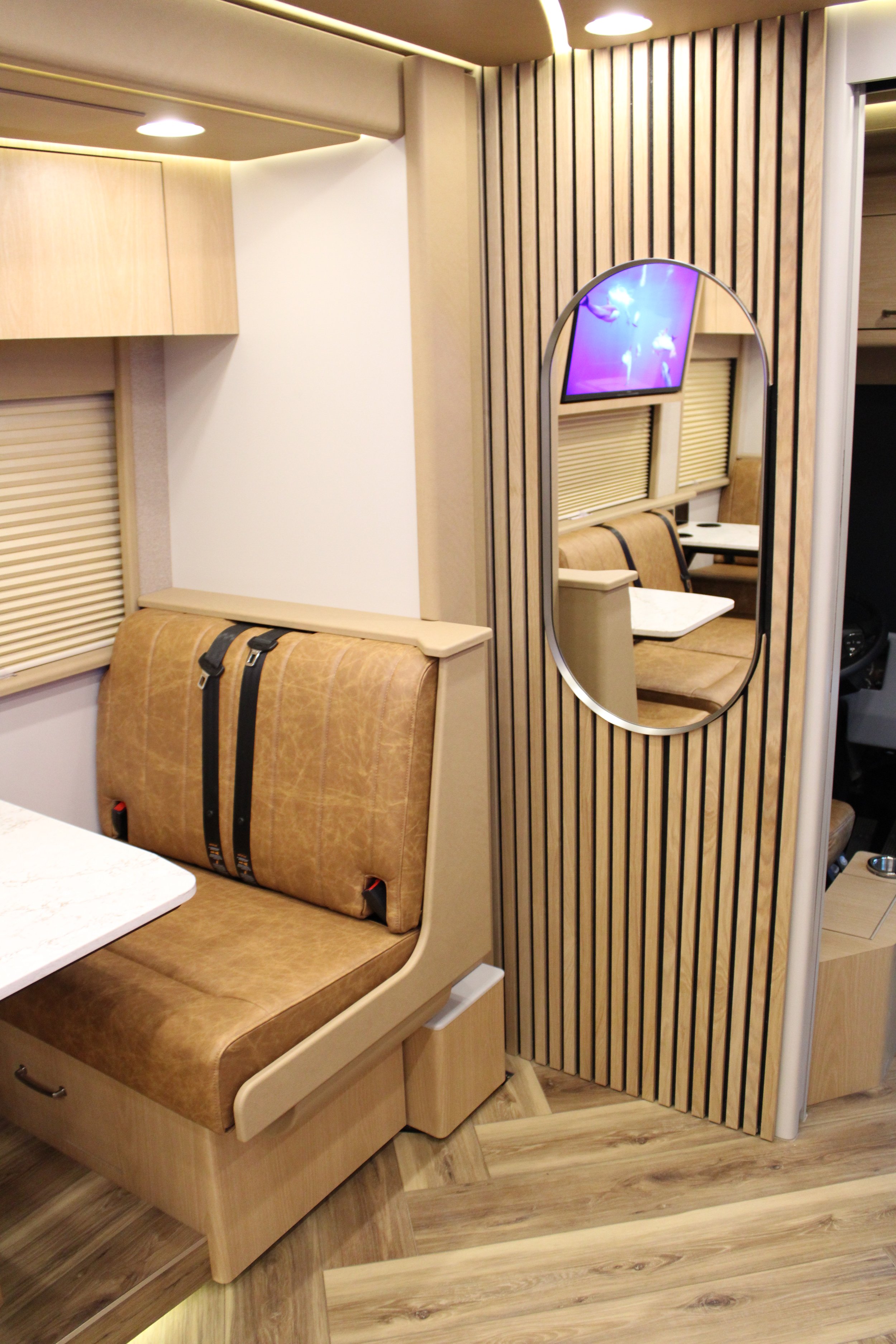 Interior of a modern RV or camper with tan leather seats, wooden paneling, a mirror on a sliding door, and a television showing an underwater scene.