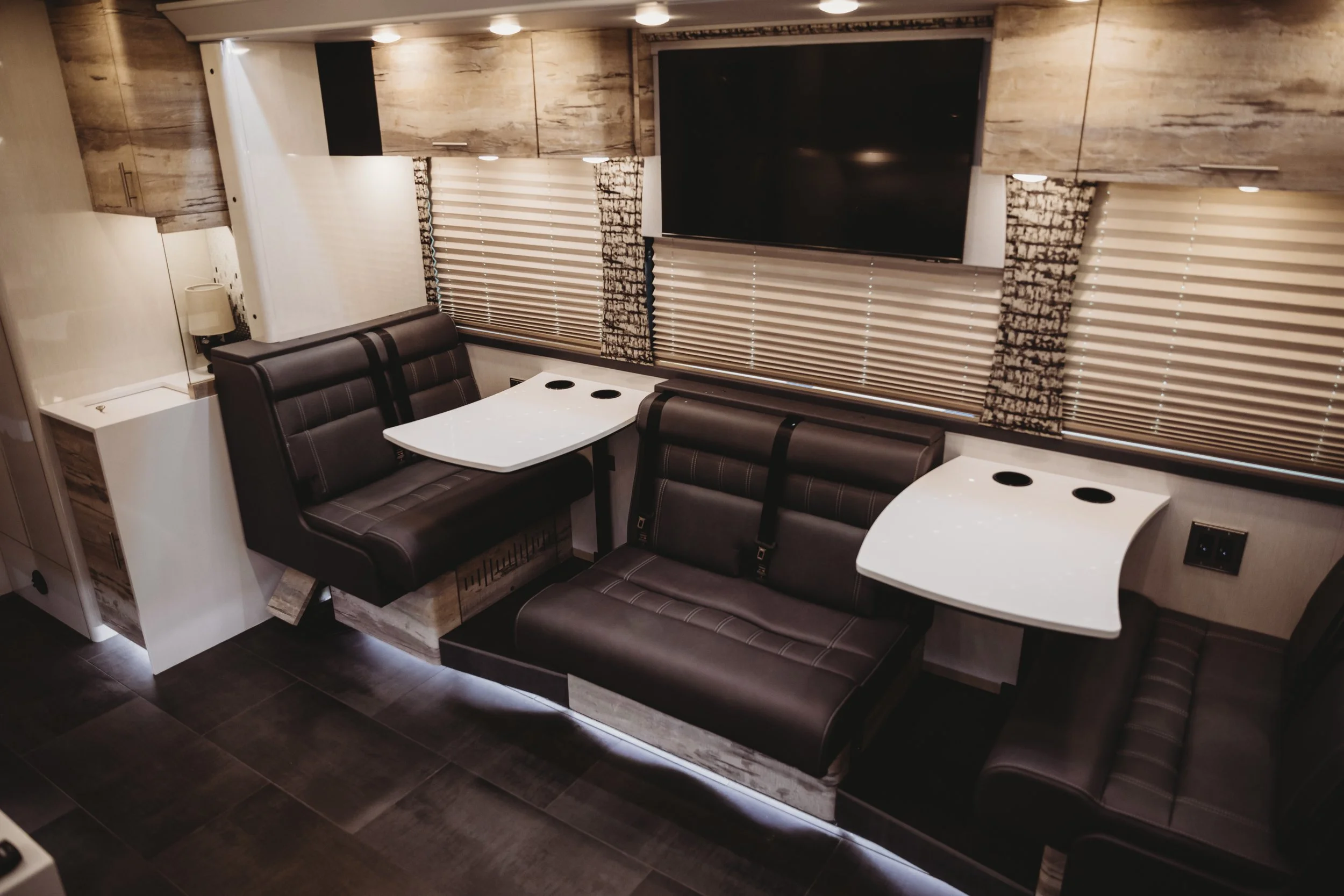 Interior of a modern RV or camper trailer with black leather seating, white plastic tables with cup holders, and a wall-mounted flat-screen TV. The space features beige and brown wood-themed decor, window blinds, and ambient lighting.