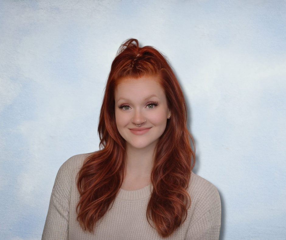 A young woman with long, wavy red hair, fair skin, and a septum piercing, smiling and wearing a beige sweater against a light blue background.
