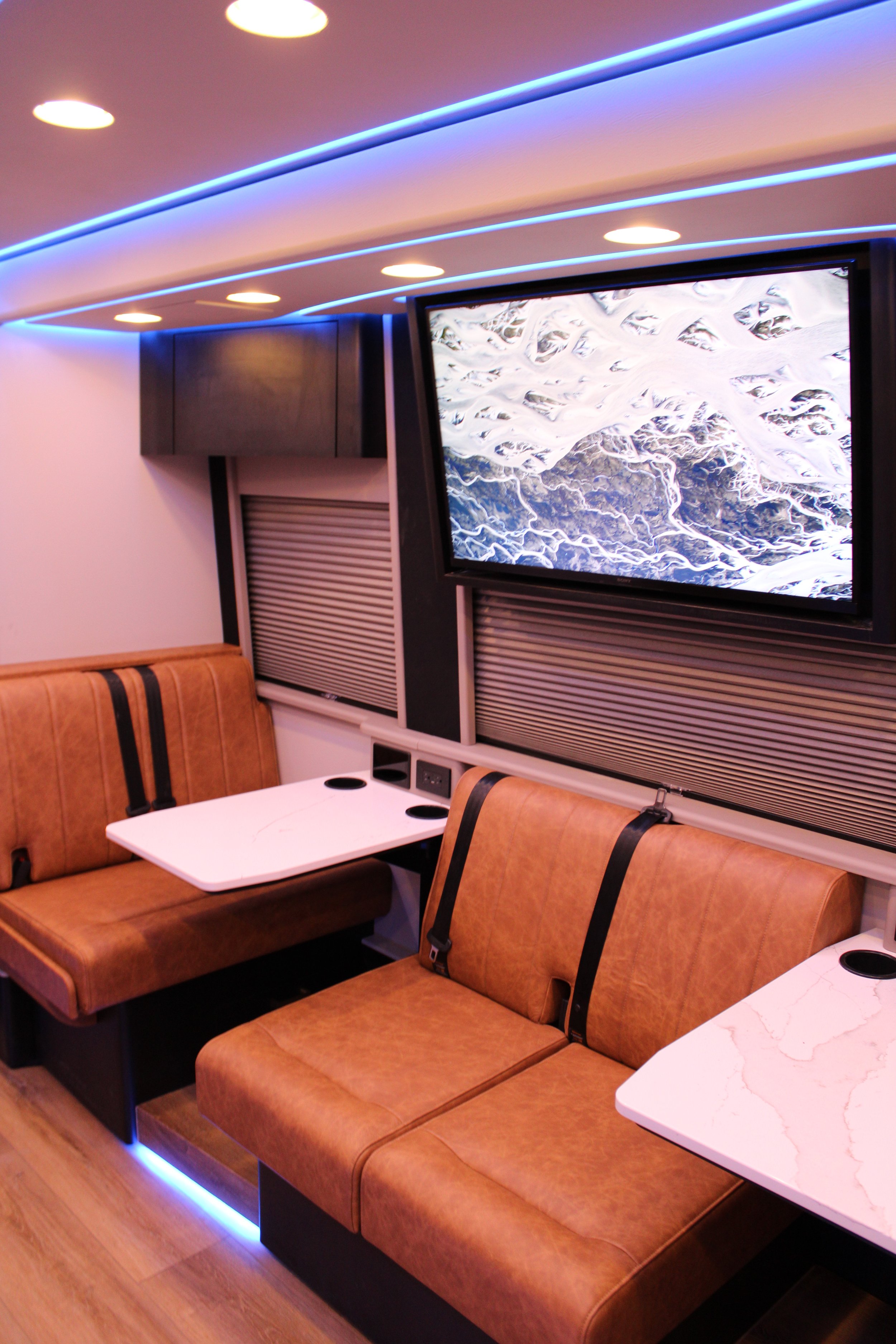 Interior of a modern lounge or recreational vehicle with brown leather seating, white marble-looking tables, a large wall-mounted TV displaying a scenic mountain view, and blue accent lighting along the ceiling.