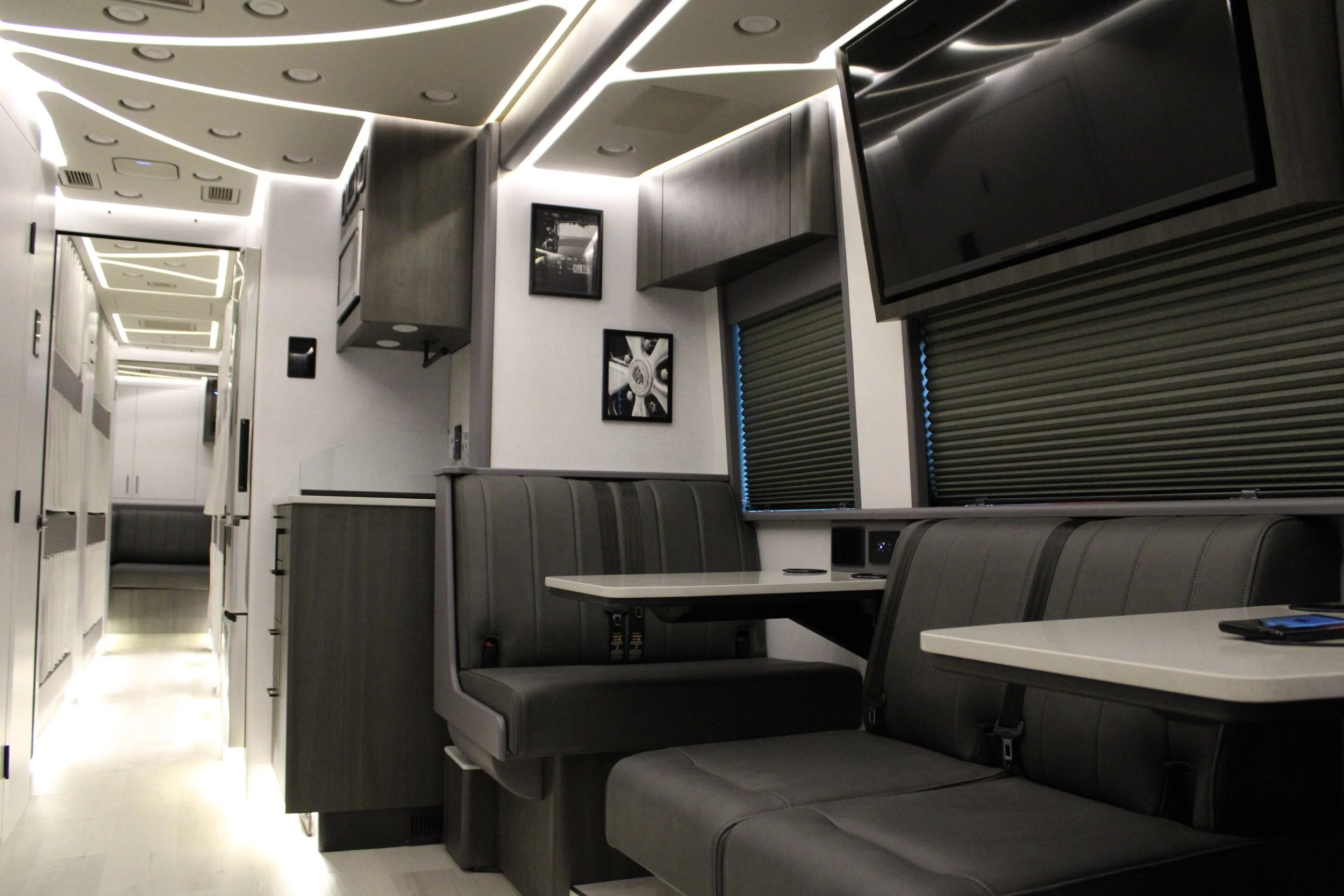 Interior of a modern, luxury RV with black leather seating, white tables, dark wood cabinets, and a large flat-screen TV.