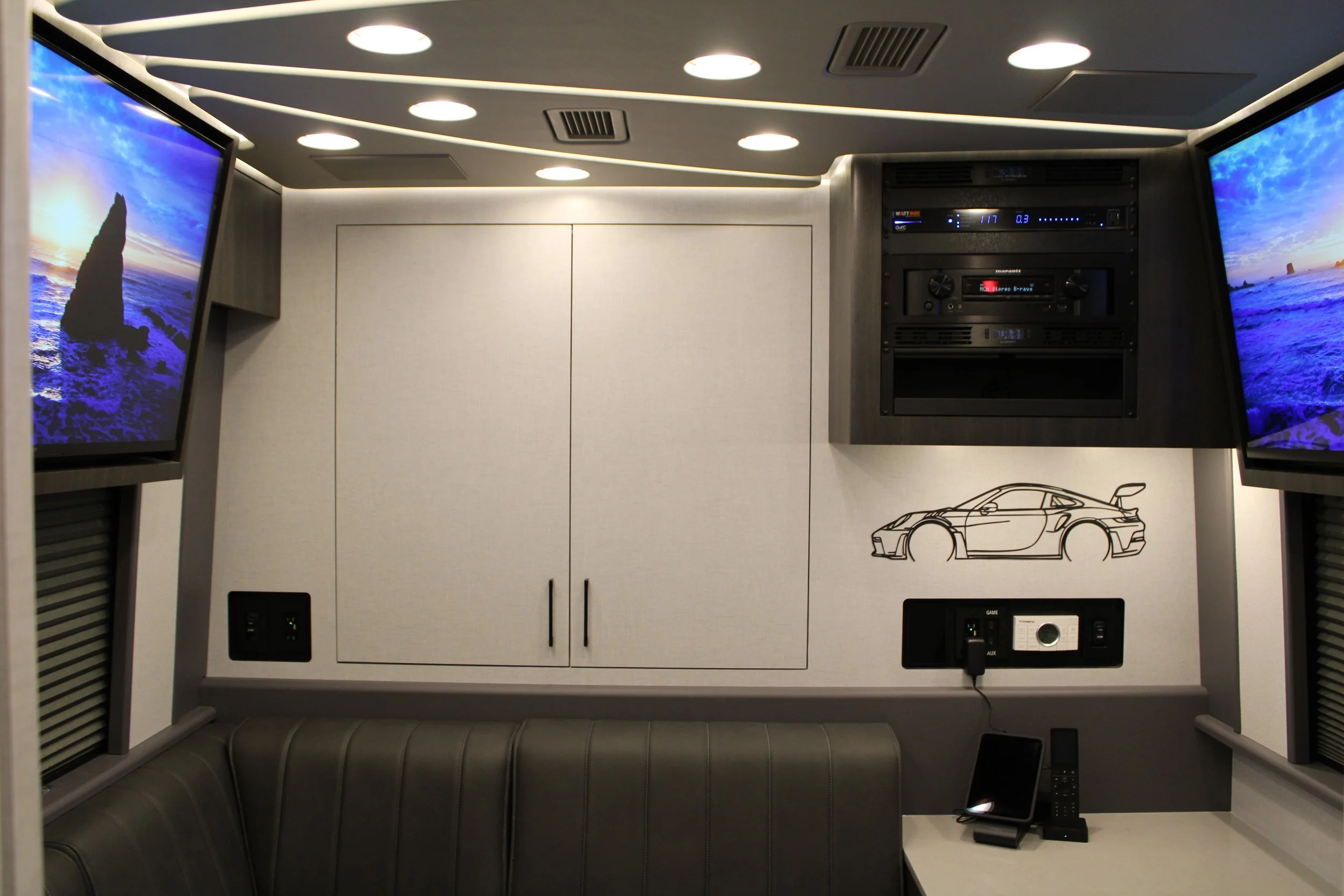 Interior of a vehicle with two wall-mounted televisions displaying ocean scenes, a drawing of a sports car on the wall, and various electronic equipment including a stereo system and a telephone.