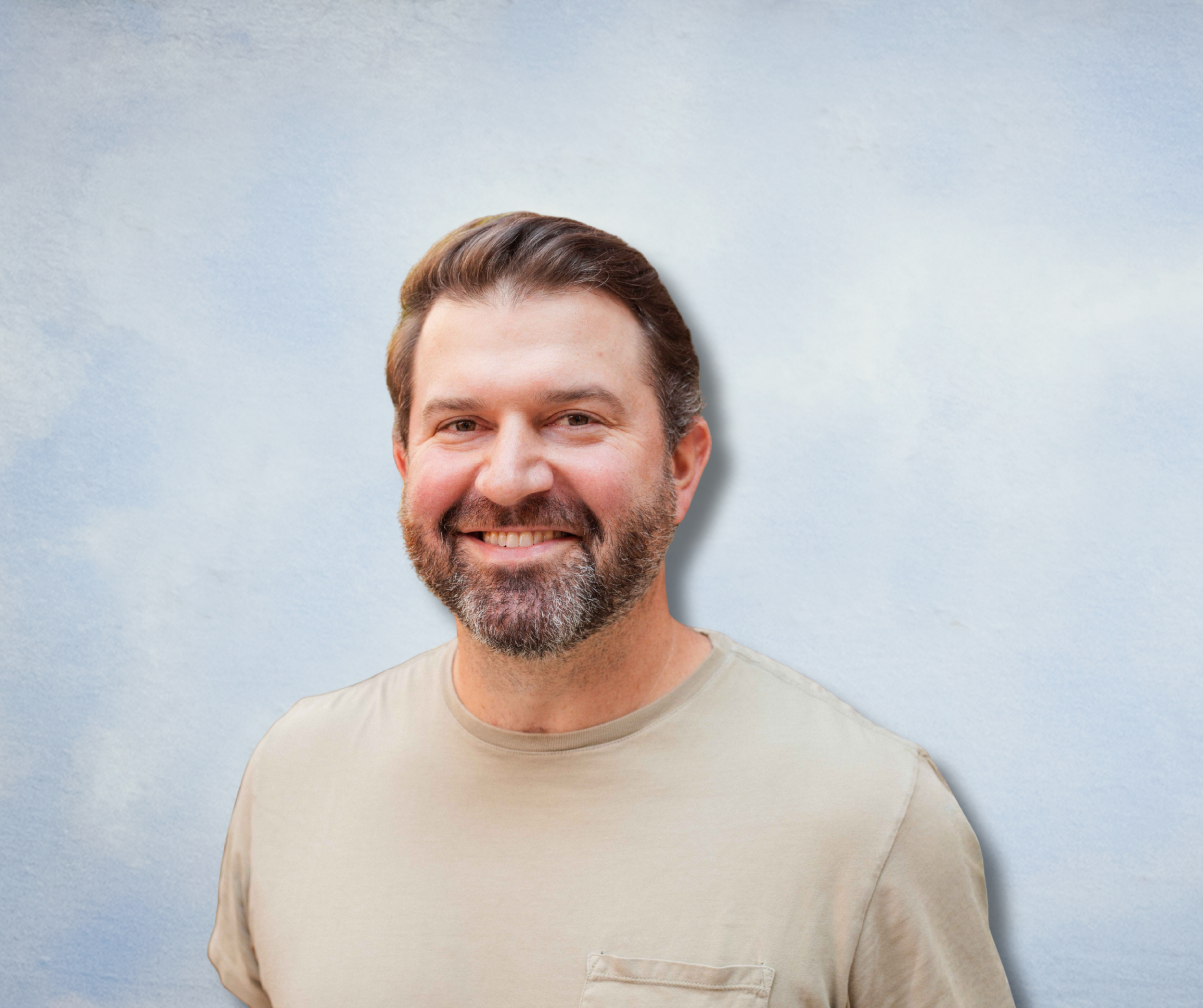A smiling man with brown hair and a beard, wearing a beige t-shirt, standing against a cloudy sky background.