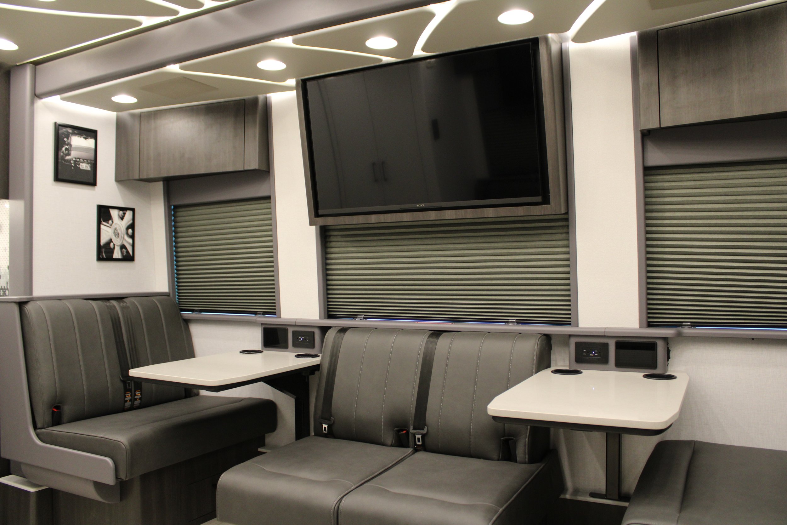 Interior of a modern train or bus with gray leather seating, two small white tables, a large flat-screen TV on the wall, and framed black and white pictures.