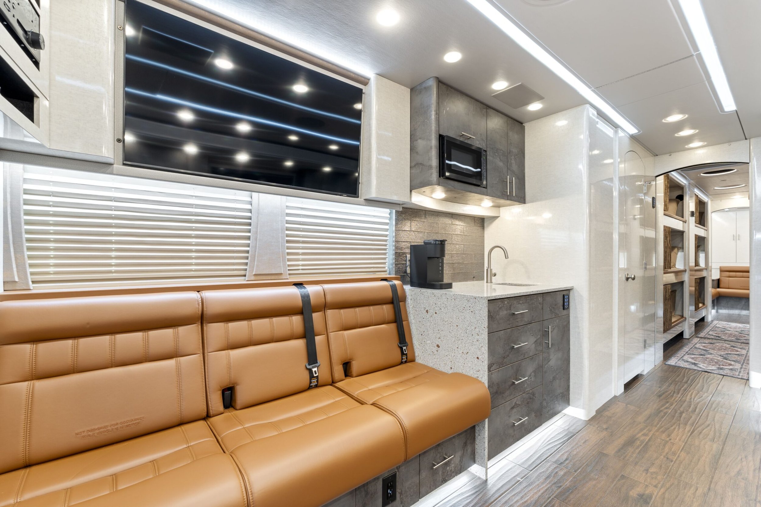 Interior of a modern office or waiting area with a tan leather bench, a kitchenette with gray cabinets, a small sink, a coffee maker, a microwave, and a large flat-screen TV mounted on the wall, with windows covered by blinds.