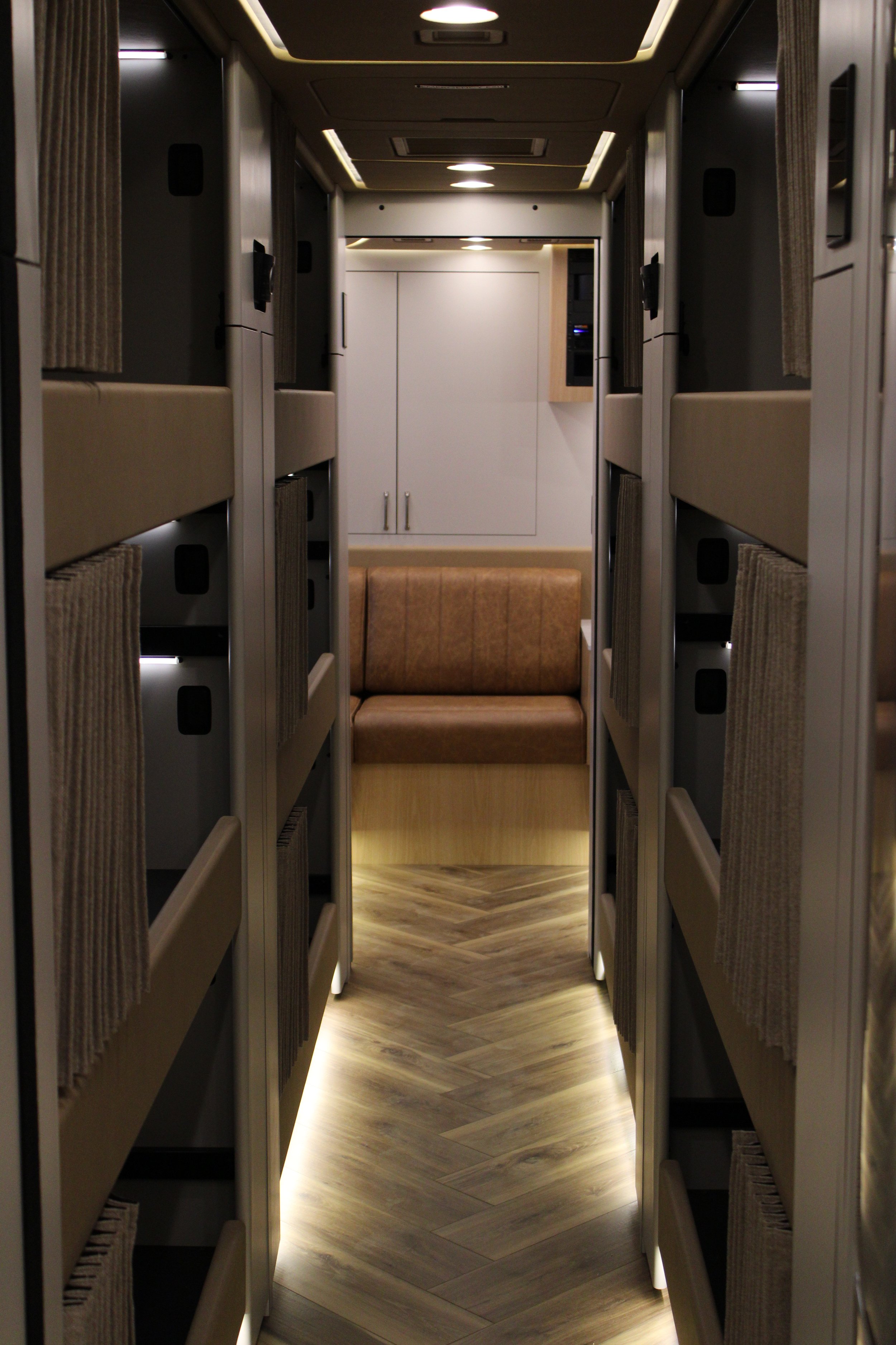 Interior of a modern airplane or train sleeping compartment with multiple bunk beds on each side, a central aisle with wood-like flooring, and a bench or seating area at the end.