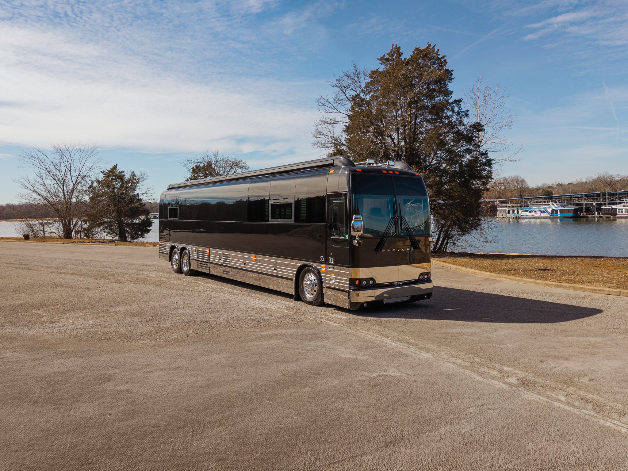 Black luxury bus parked by the water with trees and boats in the background under a blue sky.
